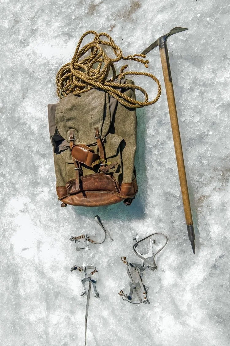 18. Vintage mountaineering gear laid out on snow, including canvas backpack, rope, ice axe, and metal crampons.jpg