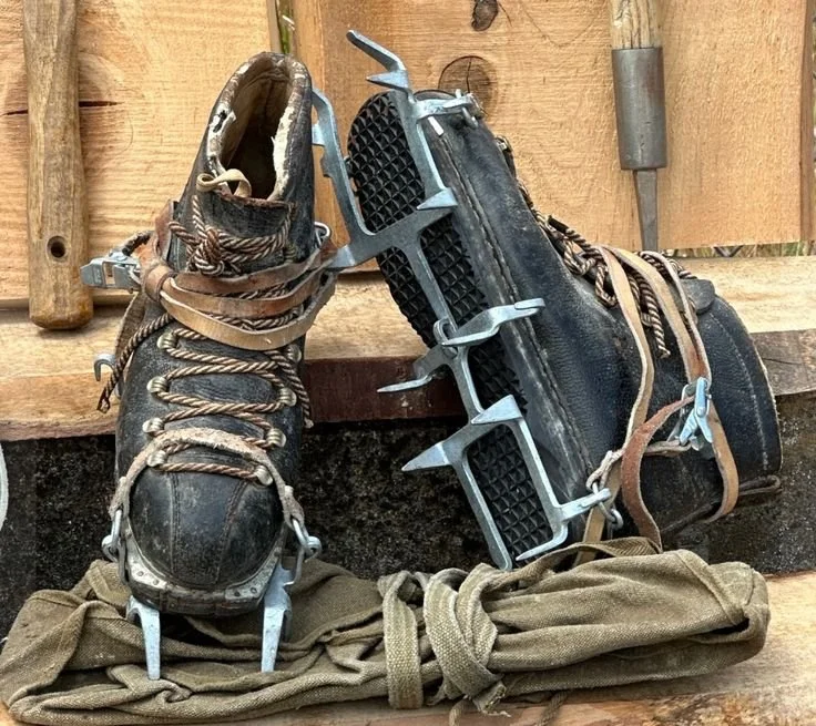 36. Vintage leather mountaineering boots fitted with metal crampons and straps, resting on a wooden surface.jpg