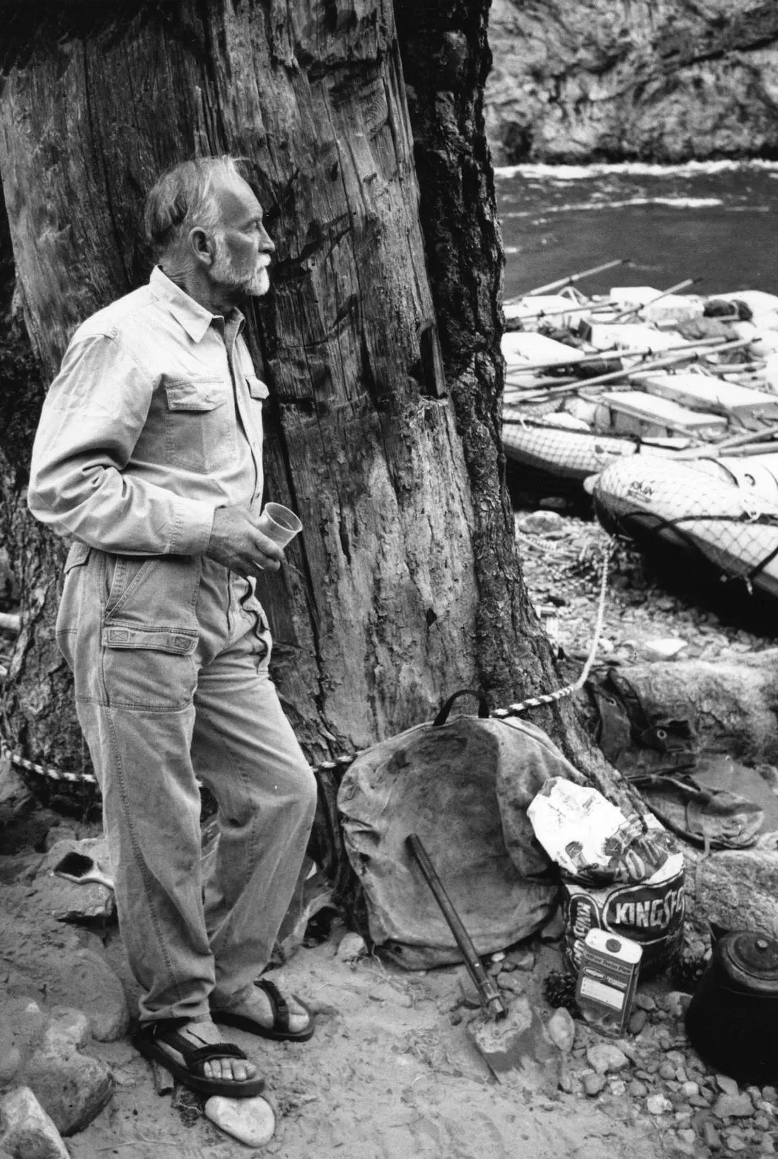 62. Black-and-white portrait of Royal Robbins standing near a campsite by a river, wearing outdoor clothing and sandals.jpg