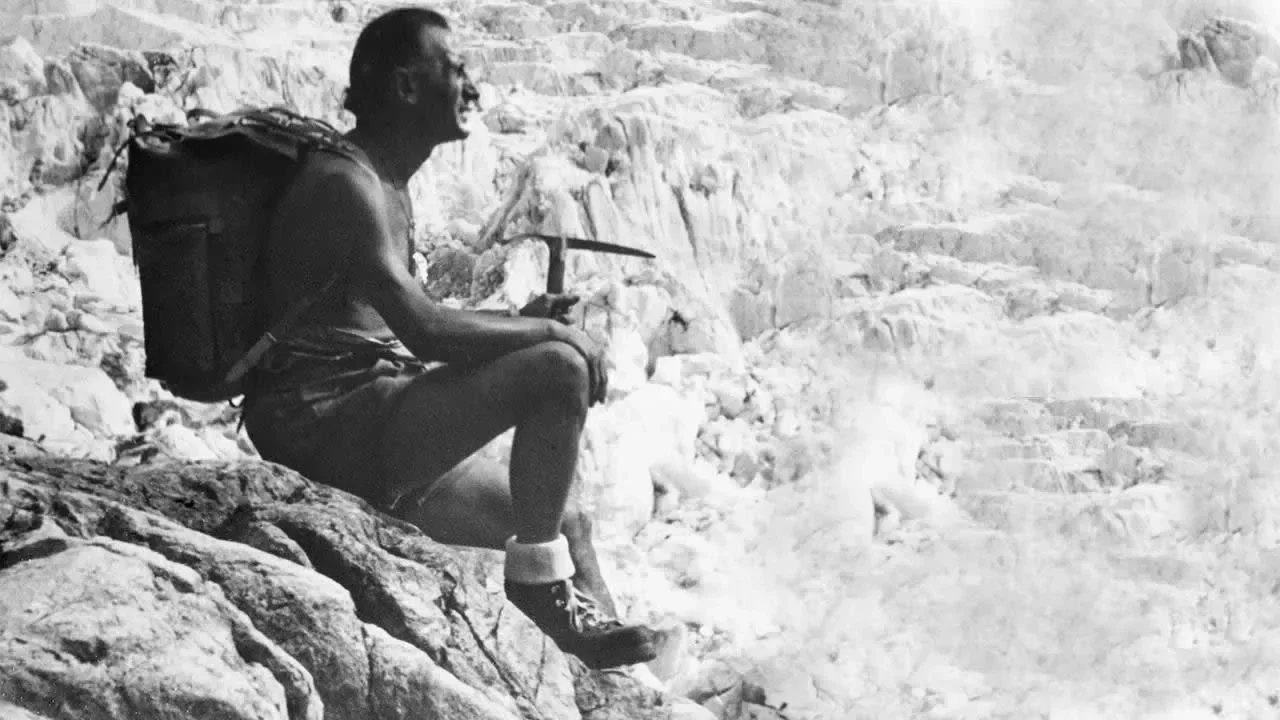 72. Black-and-white photograph of a Vitale Bramani resting on rock with ice axe and backpack in a glaciated alpine setting..jpg