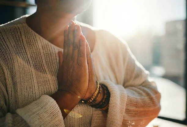 Person with hands pressed together in a prayer or meditation pose, wearing a cream sweater and multiple bracelets.