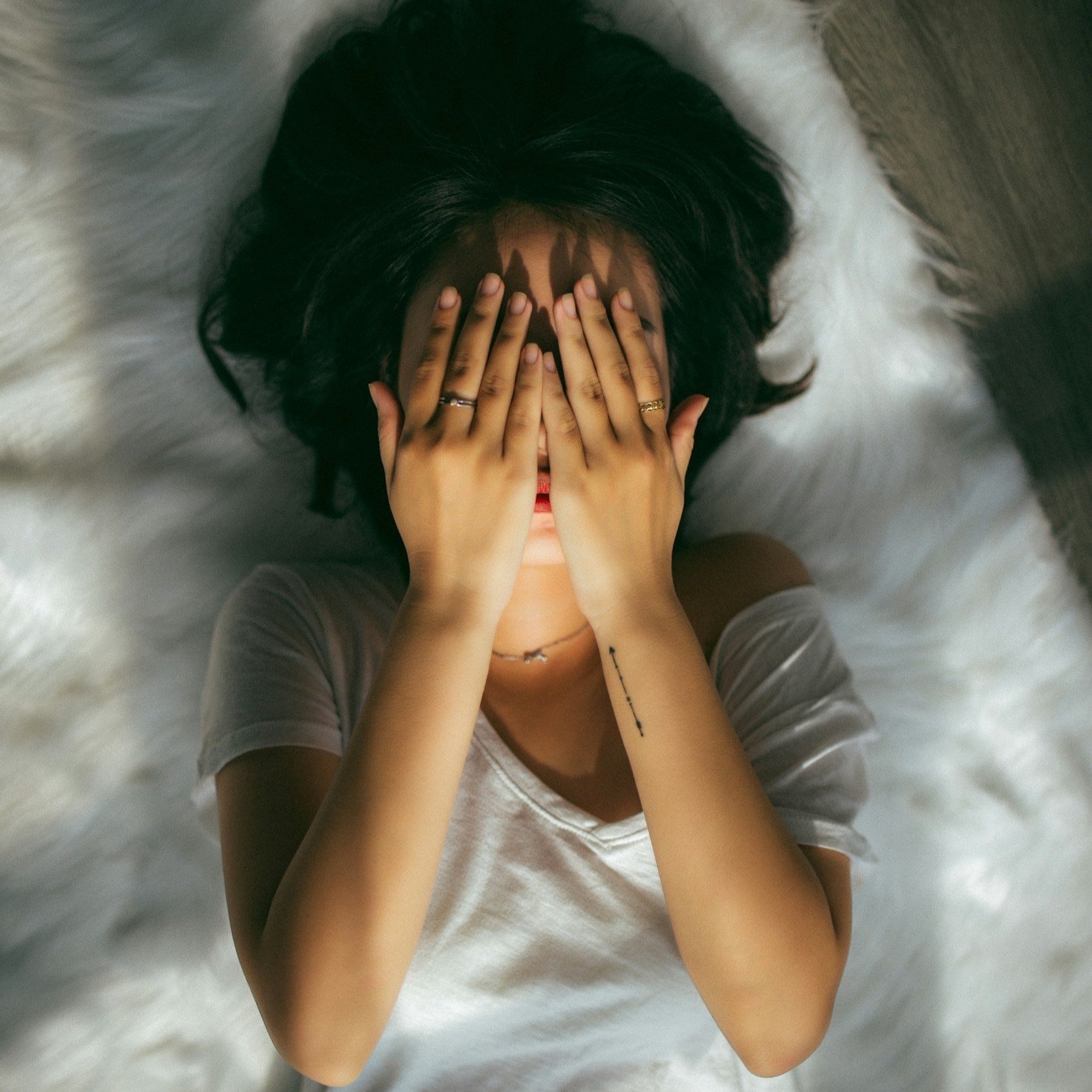 A woman with dark hair lying on a fluffy white rug, covering her face with her hands, wearing rings and a necklace.