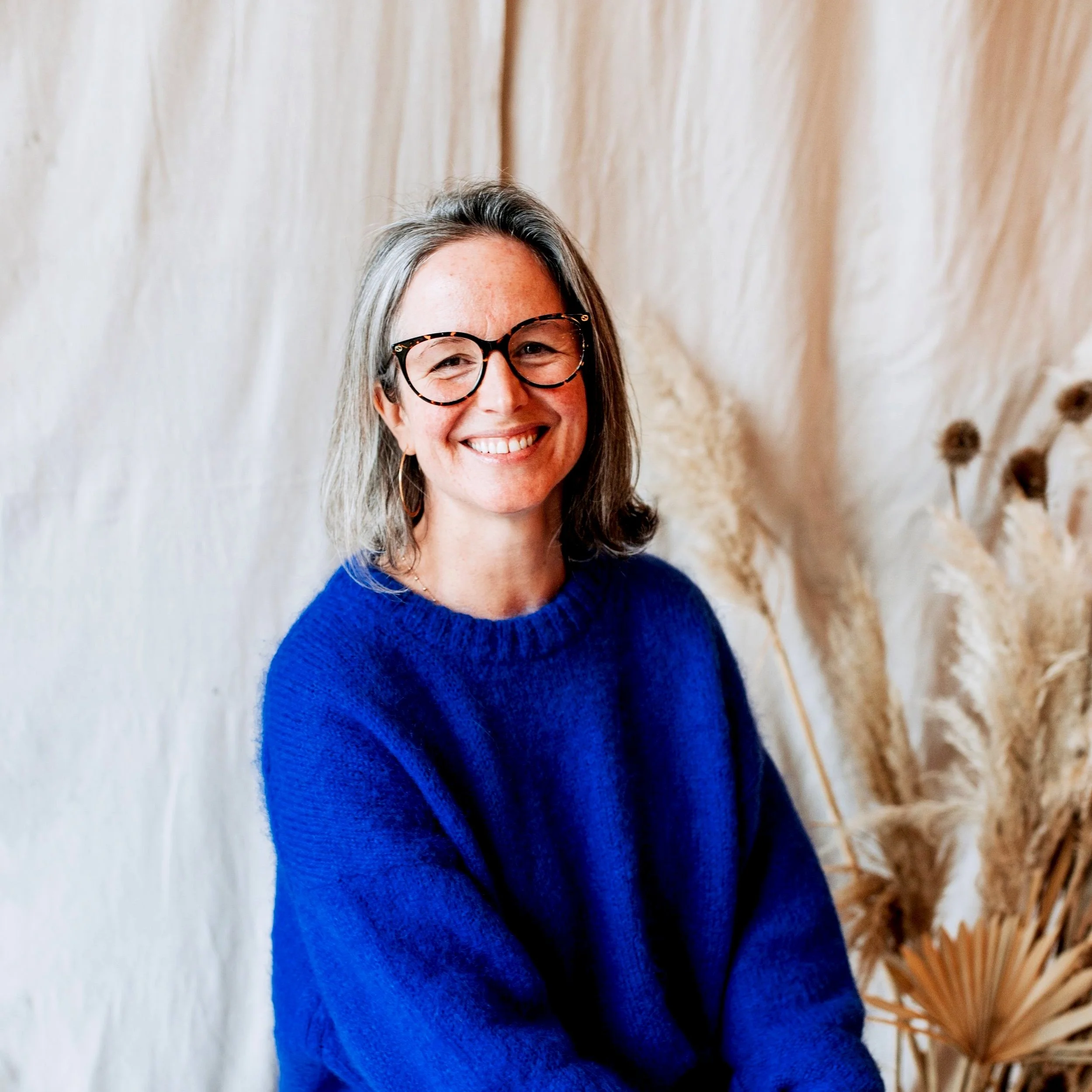 Smiling woman with shoulder-length gray hair, wearing glasses and a bright blue sweater, sitting in front of beige textured backdrop with dried plants.