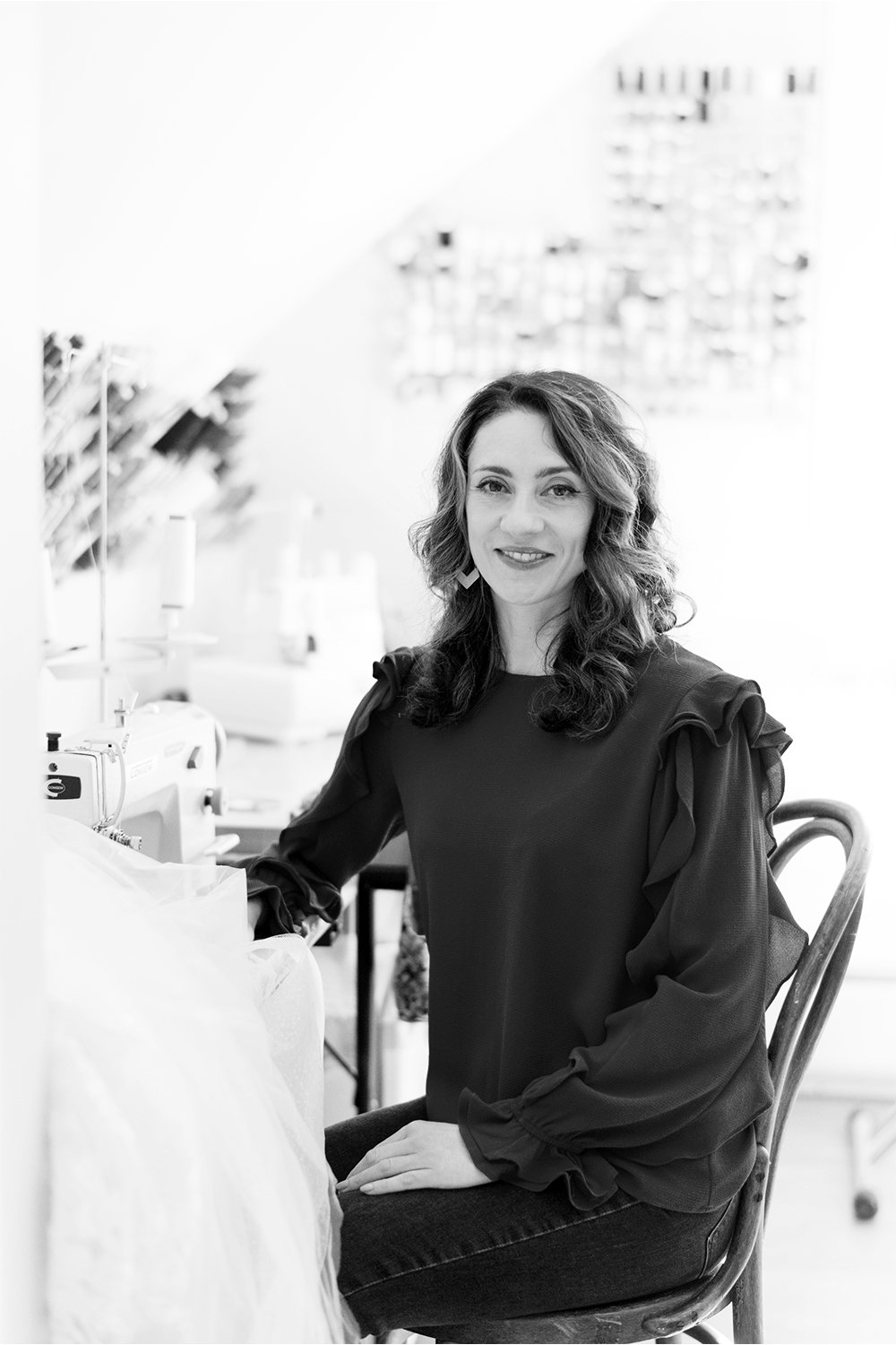 A woman with wavy hair smiling, sitting in a chair in a well-lit tailoring studio with sewing machine in background