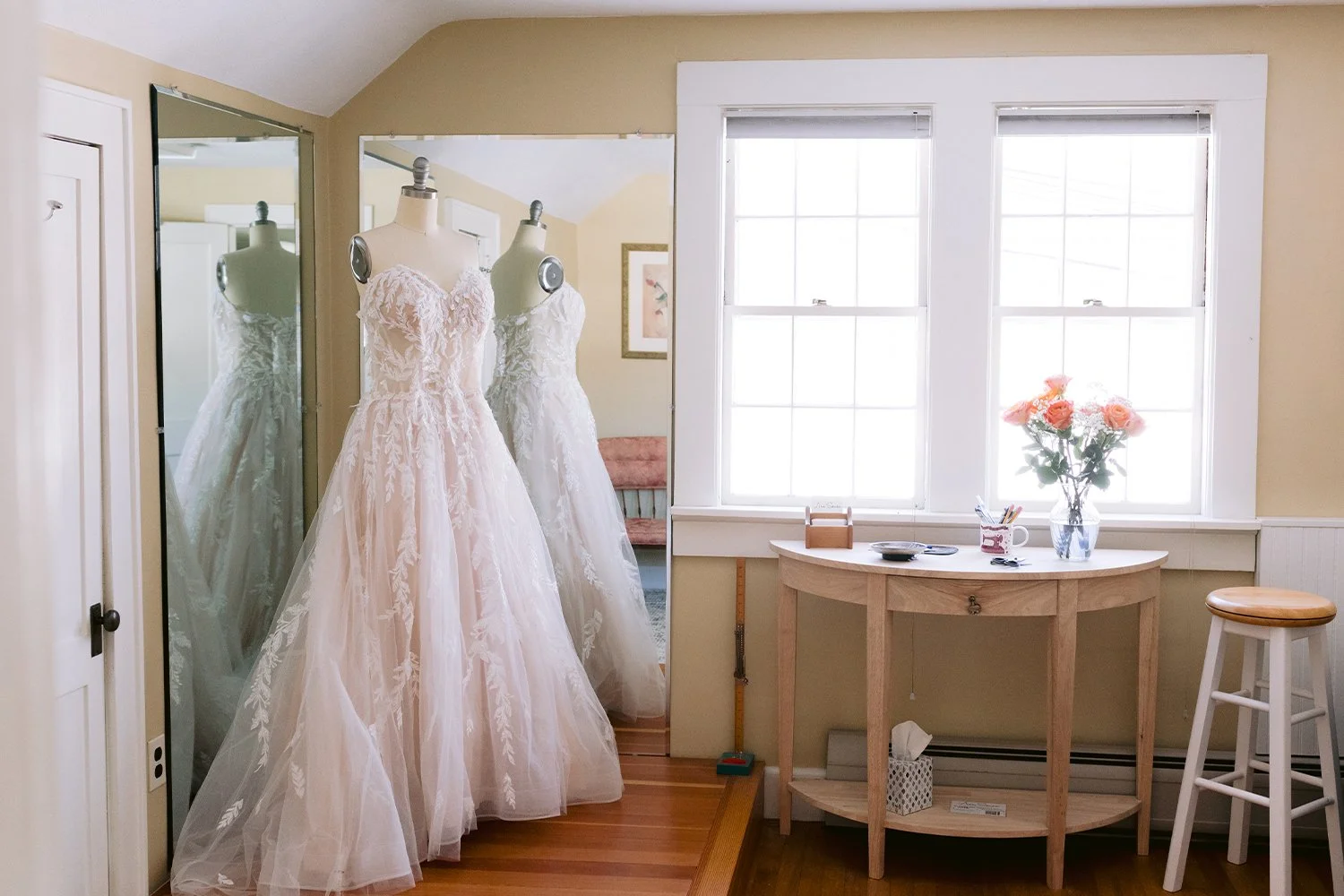 Two mannequins dressed in wedding gowns in front of a large mirror in a bridal boutique, with a window and a small table with a vase of pink roses.
