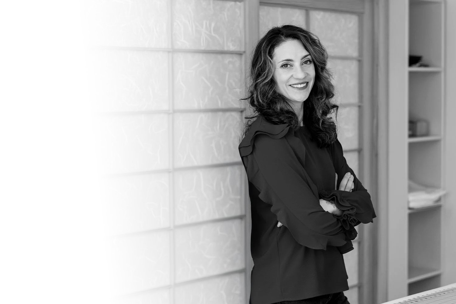 Black and white photo of a woman, Lauren Gottschalk, with curly hair, smiling, standing with her arms crossed, in an indoor setting with shelves in the background.