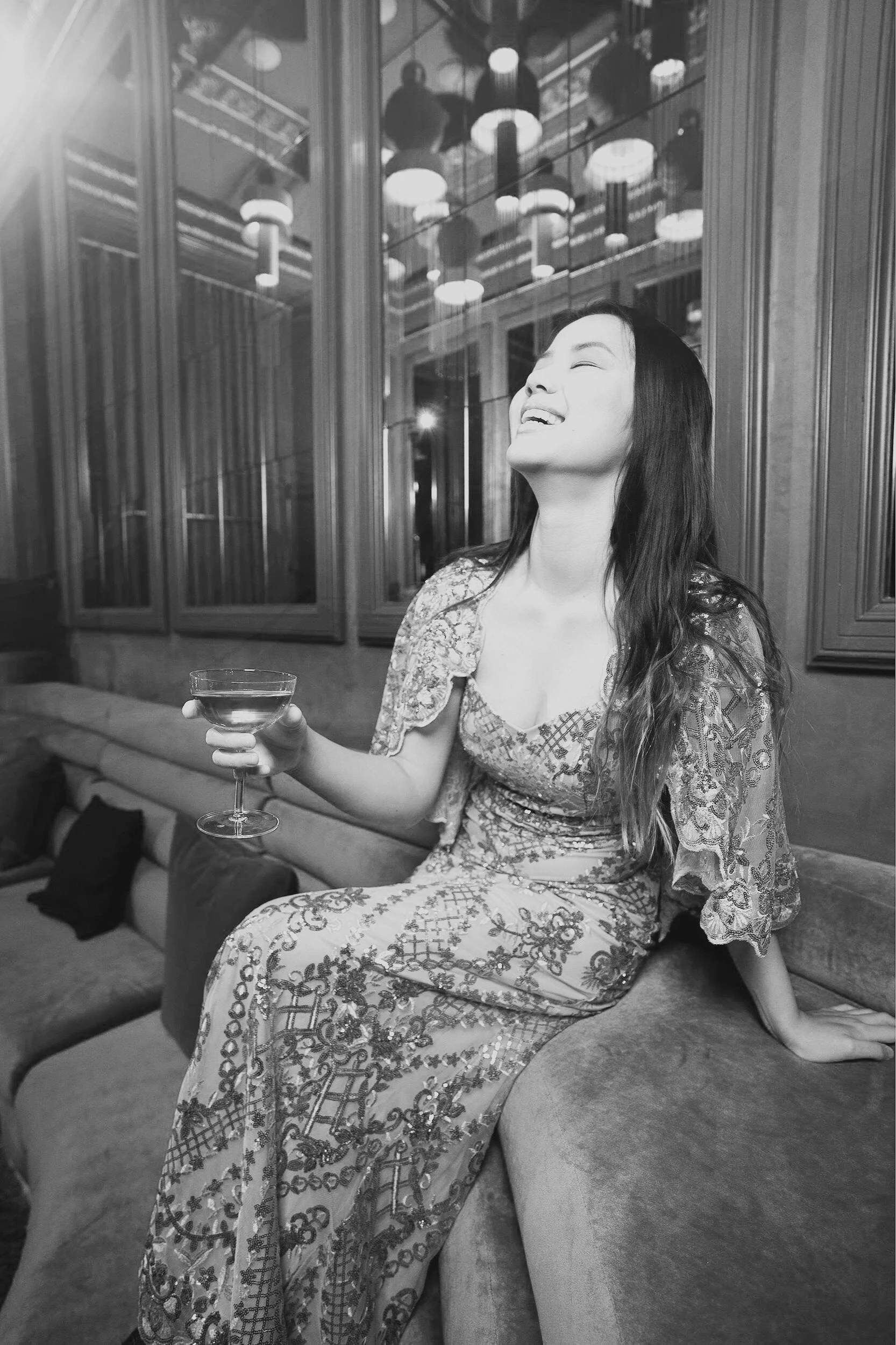 A young Asian woman, radiant in a simply stunning gown, smiles and laughs while holding an artisanal cocktail. Her designer gown, altered to perfection, features beautiful sequins and elegant scallop-edge sleeves, showcasing a blend of style, joy, an