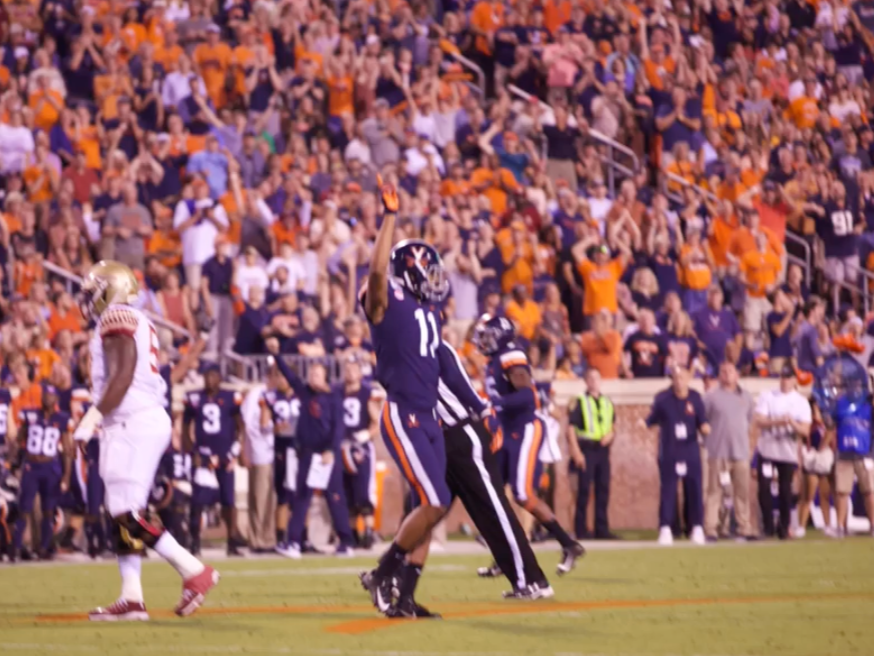 A UVA football player raises his arms in celebration on the field at Scott Stadium as the crowd cheers in the background during a night game.