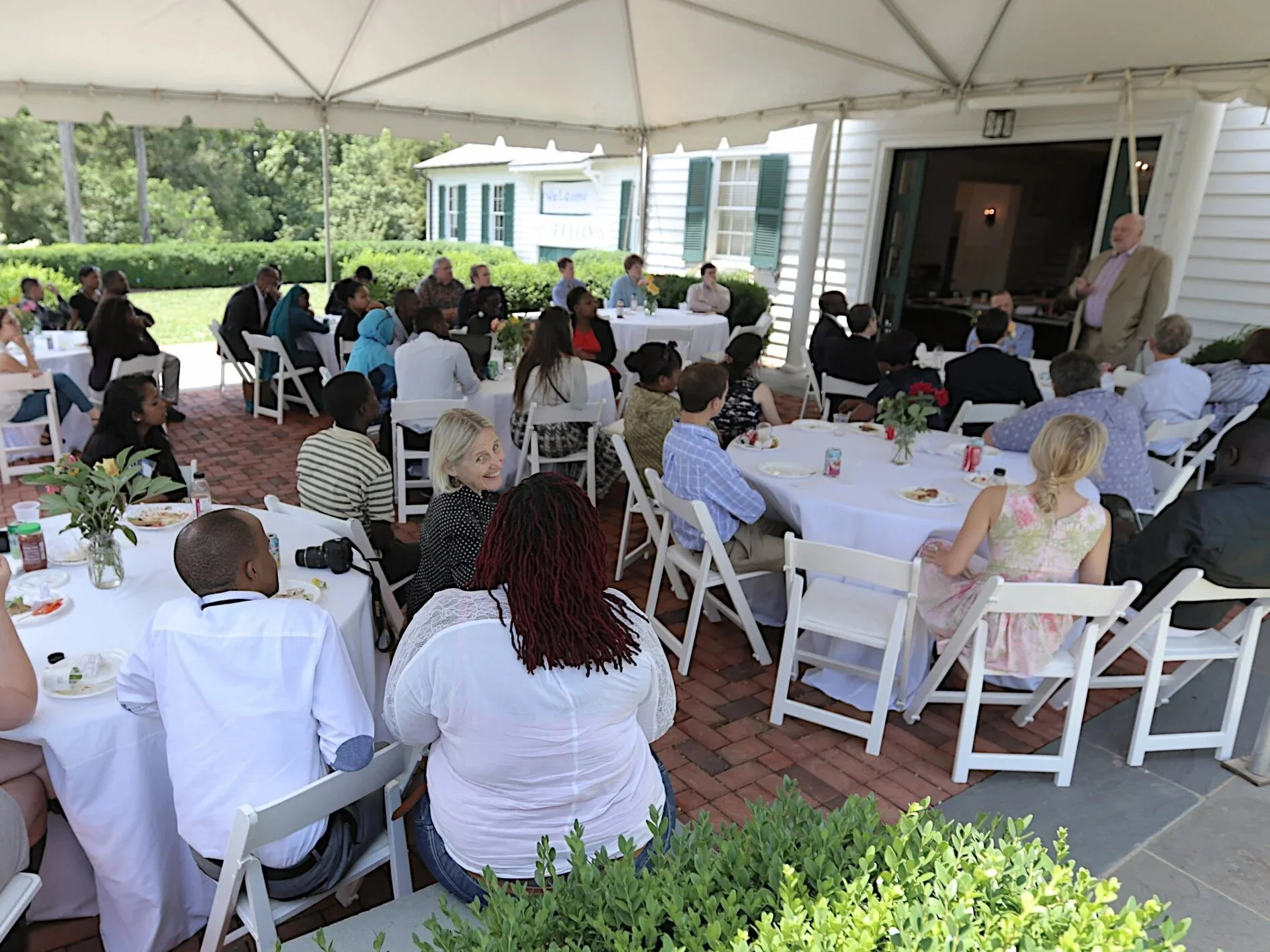 An event at the Morven Meeting barn, taking place on the front patio under a white tent. Guests sit around round tables covered in white table cloths, listening to a speaker who stands at the Meeting Barn sliding doors.