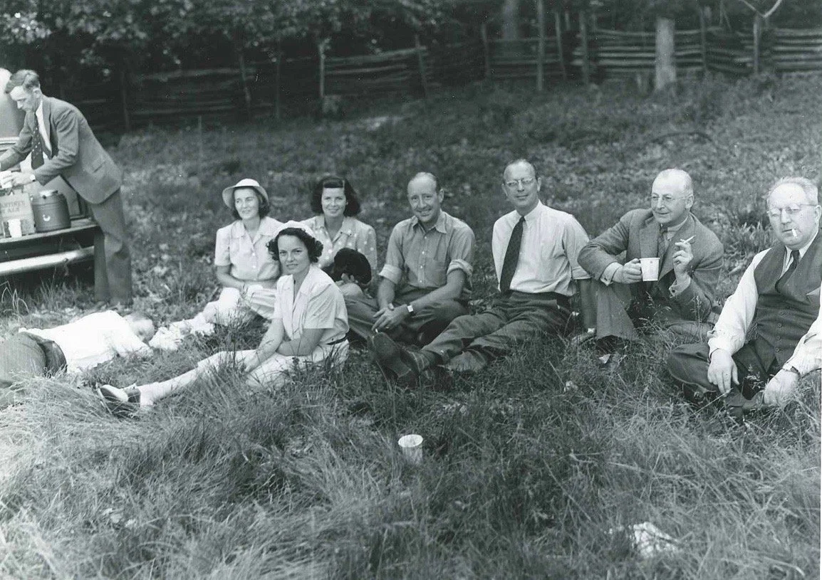 The Stone family sits in a grassy field, with Curtis Thacker at the truck getting a drink, the background shows a fence line against a tree lined edge.