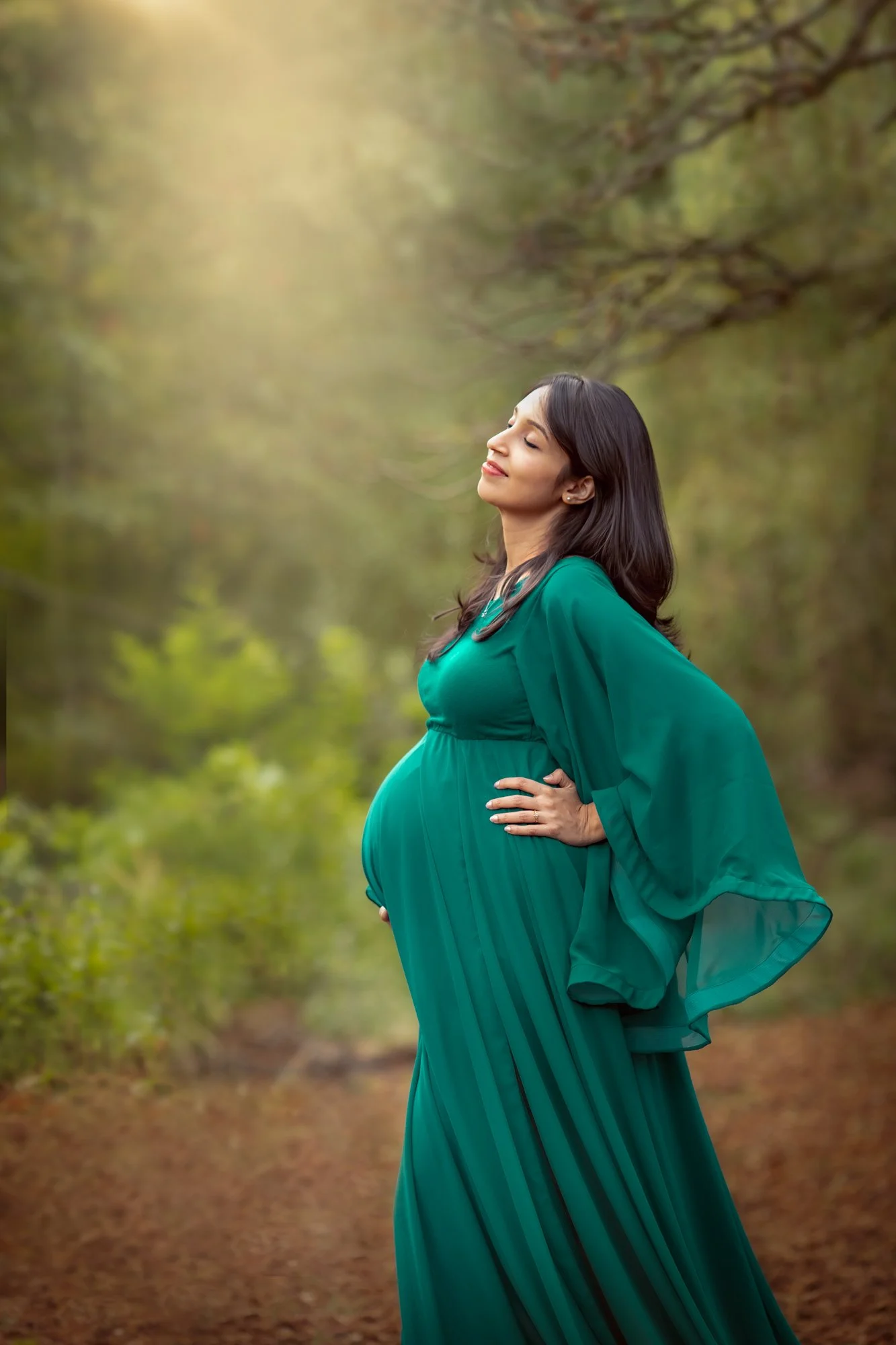 A pregnant woman in a green dress standing outdoors on a trail surrounded by trees with her eyes closed, smiling, and resting her hand on her hip.