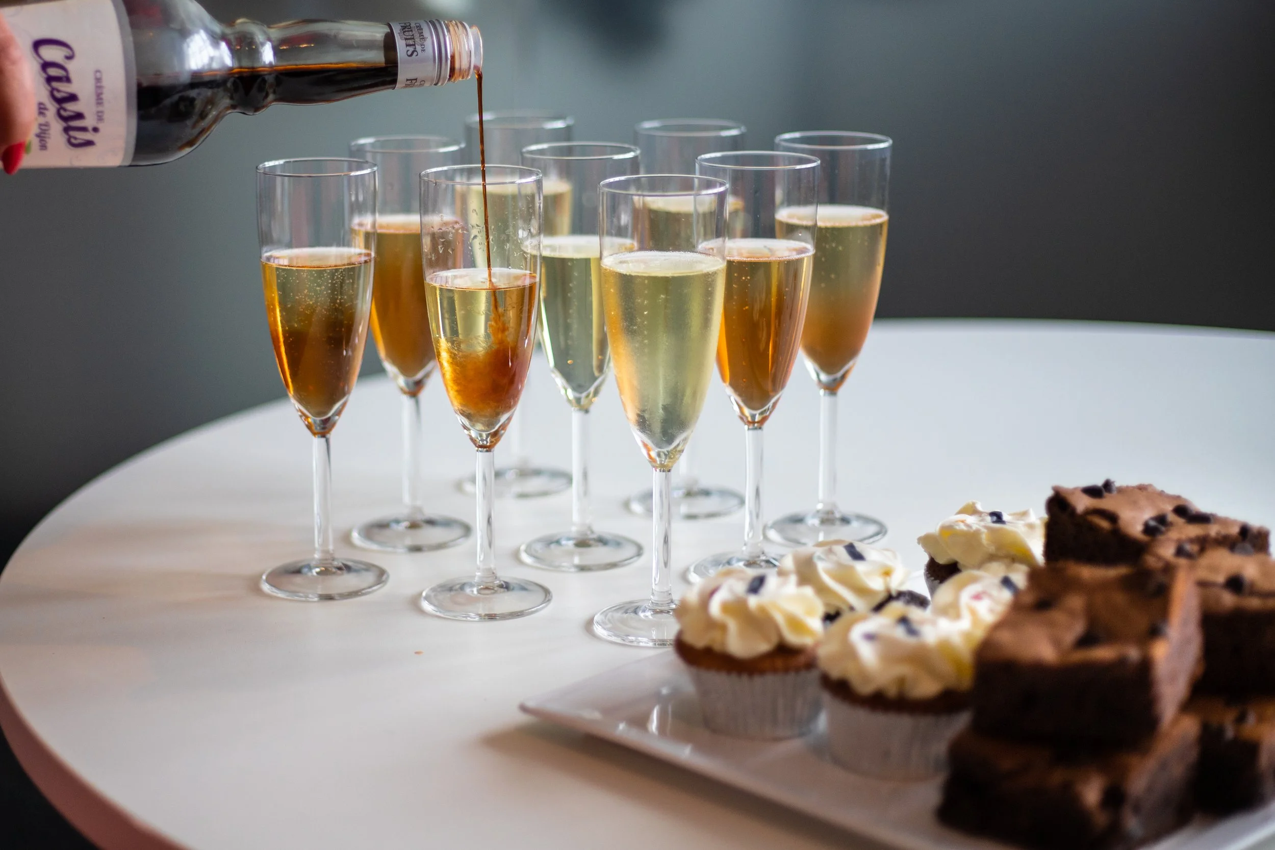 A tray of champagne glasses filled with sparkling wine, some in the process of being poured from a bottle in the background, with assorted desserts in front, including cupcakes with frosting and chocolate chip cookies.