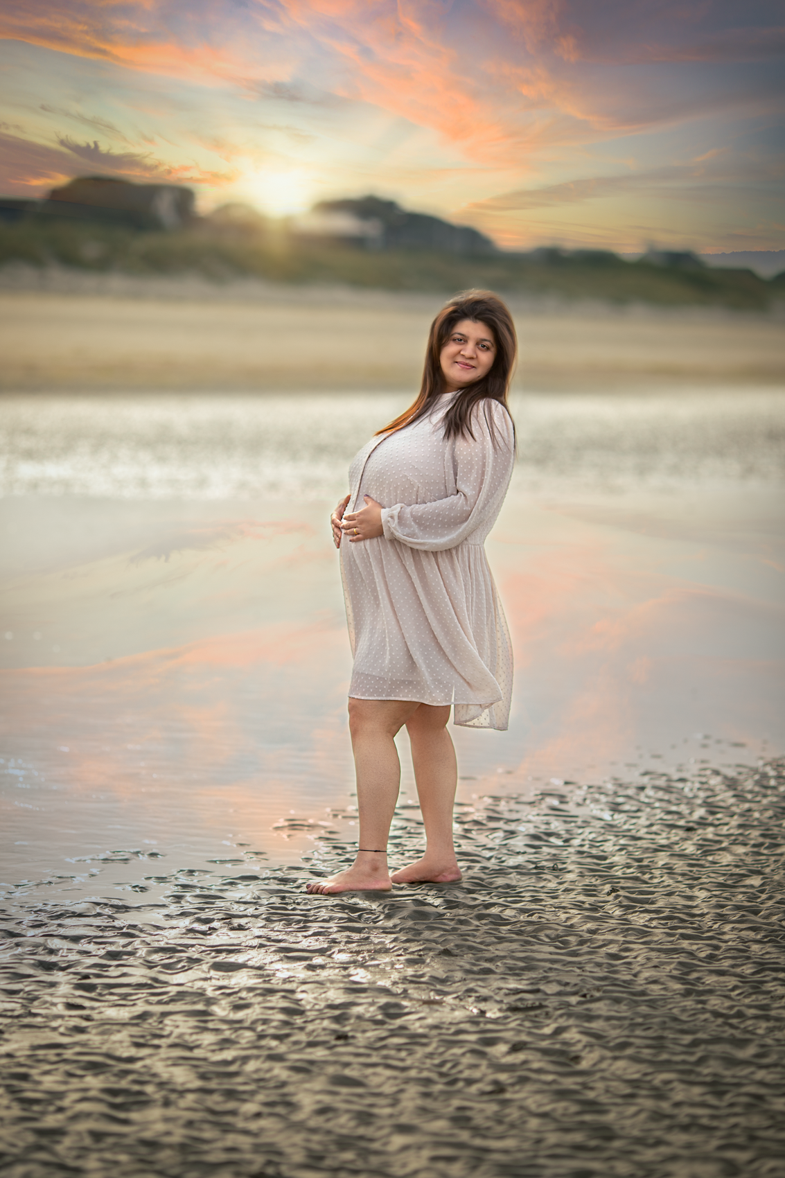 Pregnant woman in a white dress standing barefoot on wet sand near water during sunset with colorful clouds.