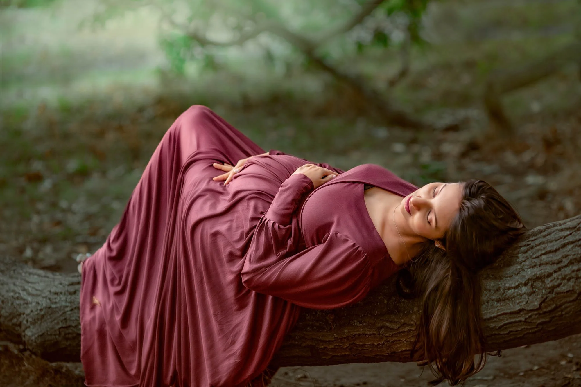 Woman in a long, burgundy dress lying on the ground with her head resting on a fallen tree trunk in a forest.