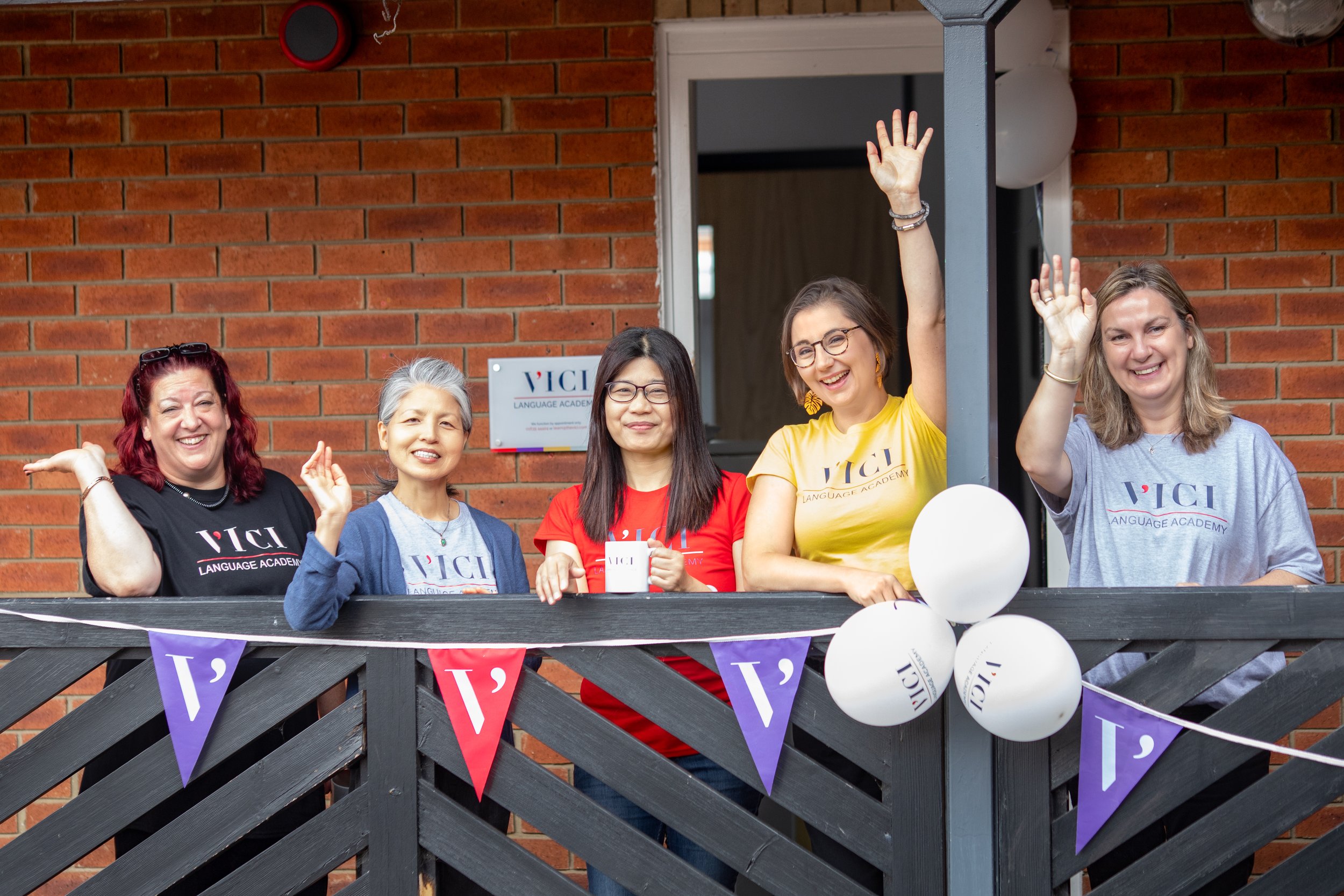 Group of five women standing on a balcony, smiling and waving, at VICI Language Academy's open event, captured by Newbury event photographer