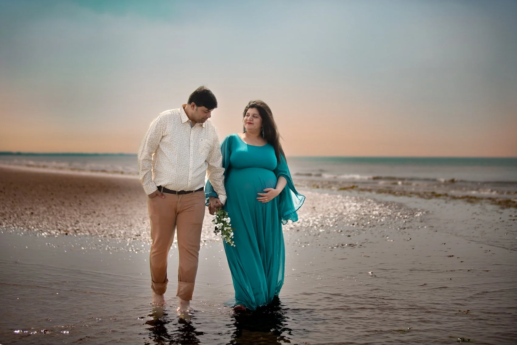 A pregnant woman in a long turquoise dress and a man in a light shirt and beige pants walking hand in hand along the beach during sunset, with the woman holding a small bouquet of flowers.