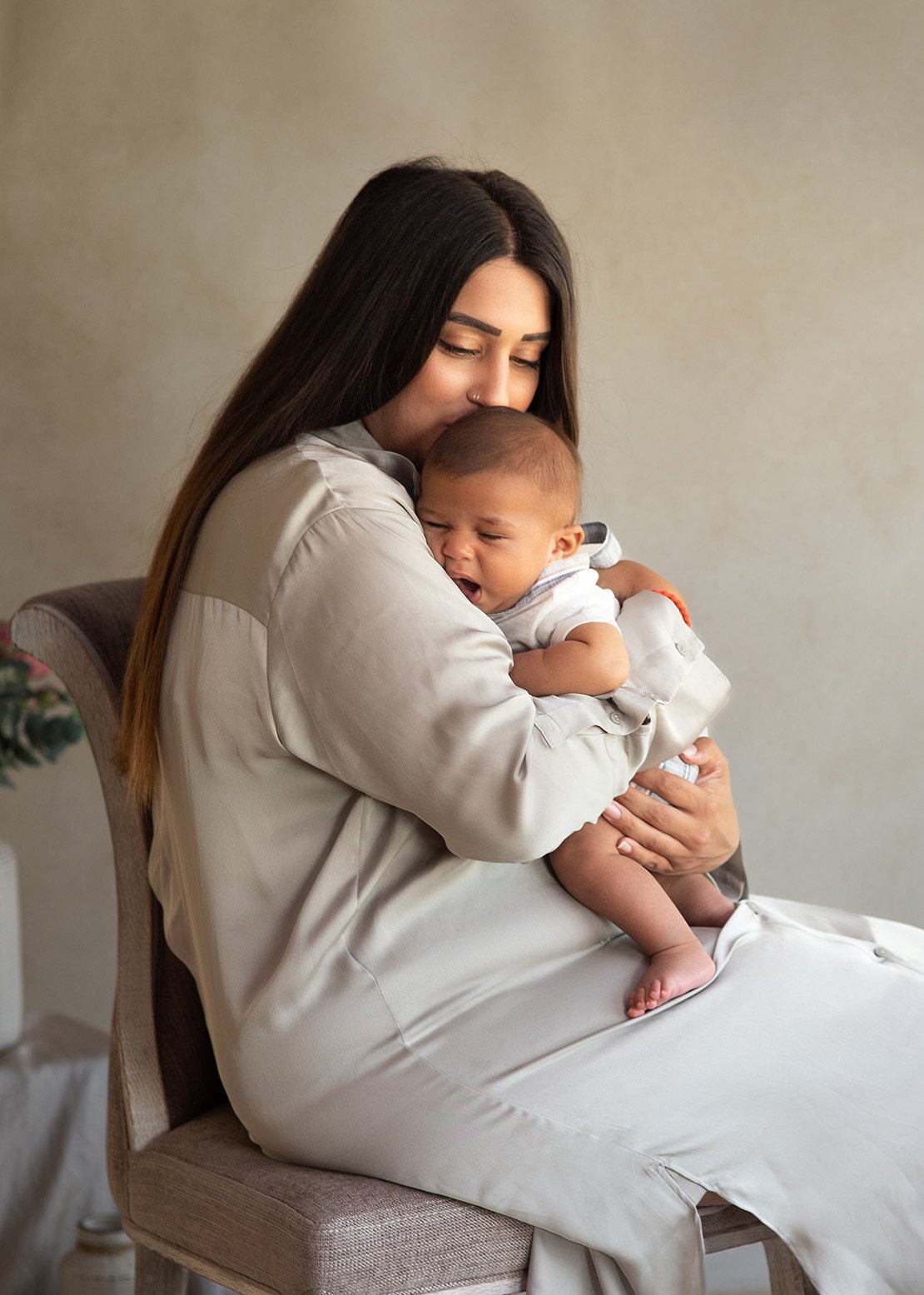 A woman holding a crying baby in a white shirt, sitting on a chair against a neutral background, caputured at newborn home photoshoot, Reading