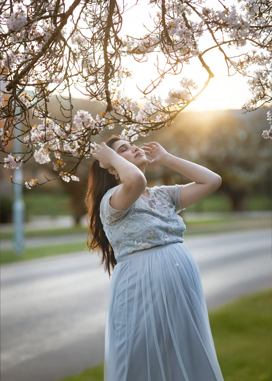 A pregnant woman in a light blue dress standing outdoors under blooming cherry blossom trees during sunset, with her eyes closed and hands gently touching her head.