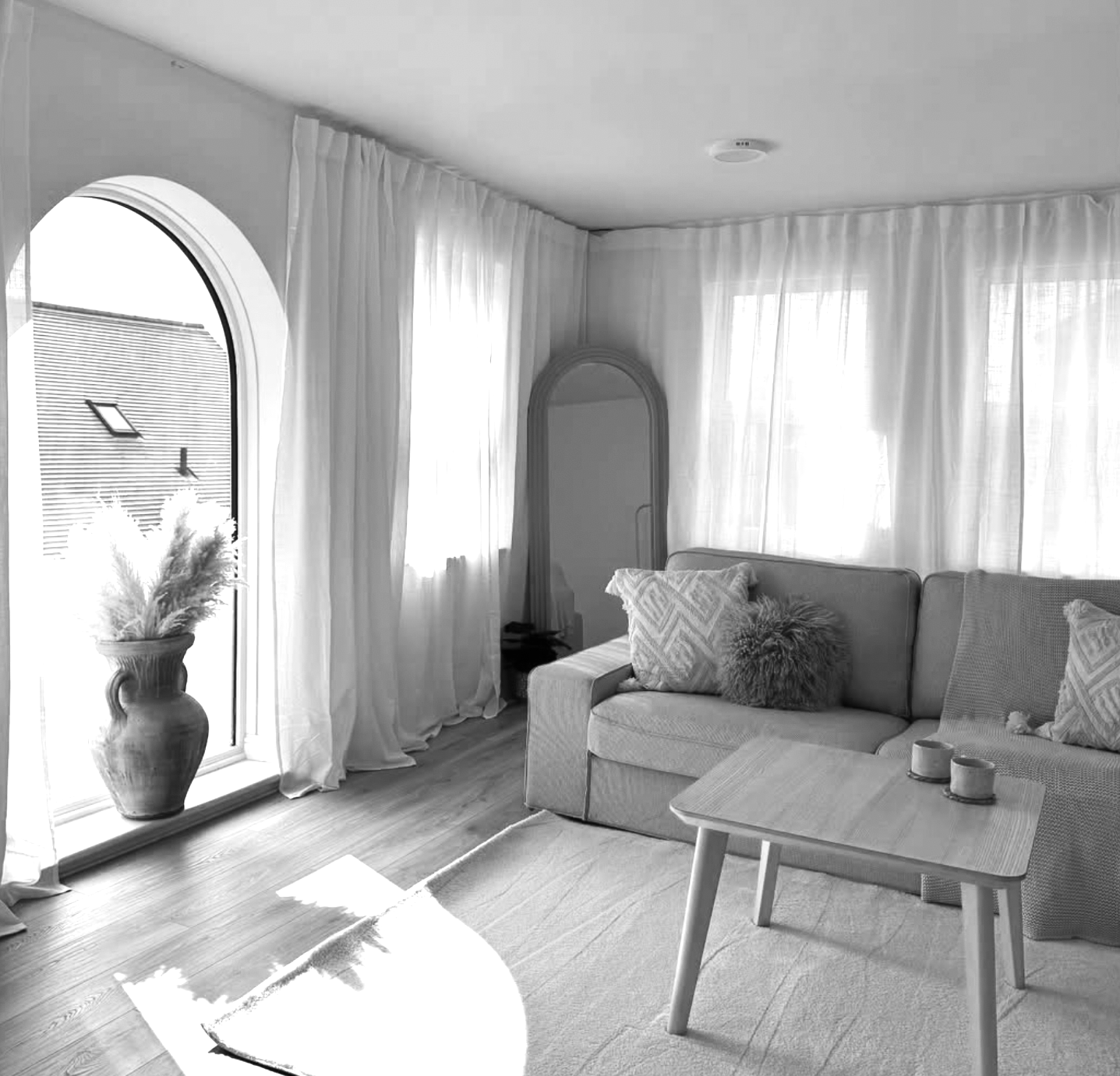 Living room with large arched window, sheer curtains, and potted plant, sofa with patterned pillows, small wooden coffee table, and standing mirror.