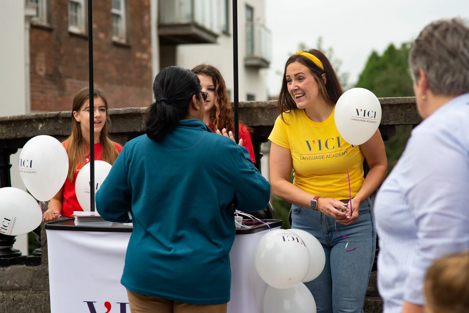 Woman wearing VICI Language Academy shirt talking to a visitor at an outdoor event booth, captured by Newbury event photographer