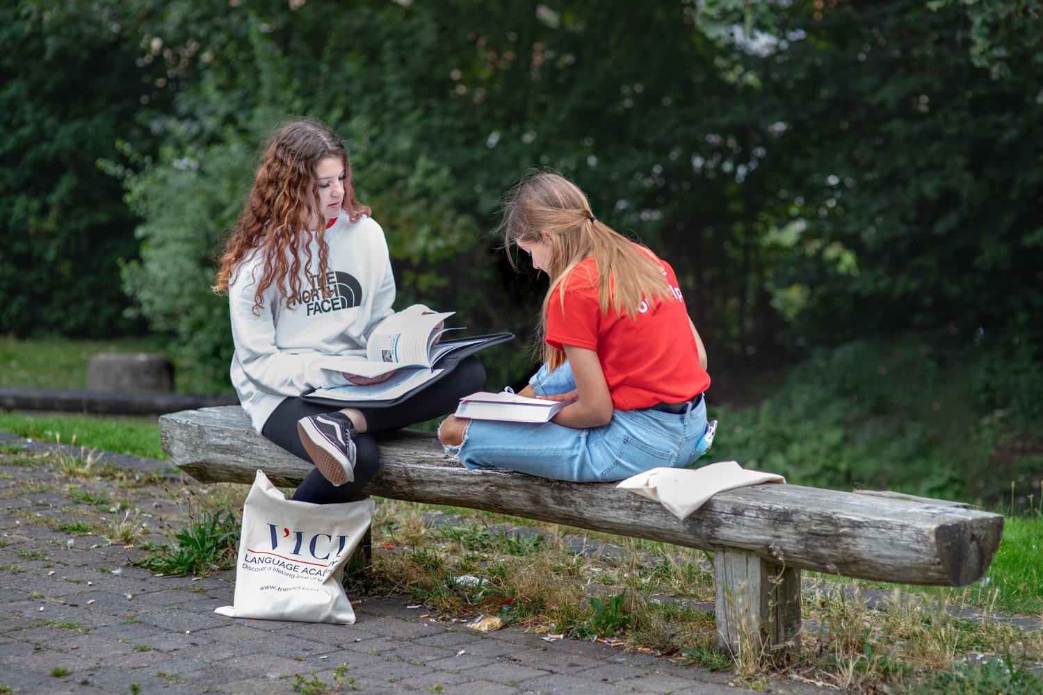 Two young girls sitting on a wooden bench outdoors reading books together, captured at branding photoshoot for VICI language academy by reading branding photographer