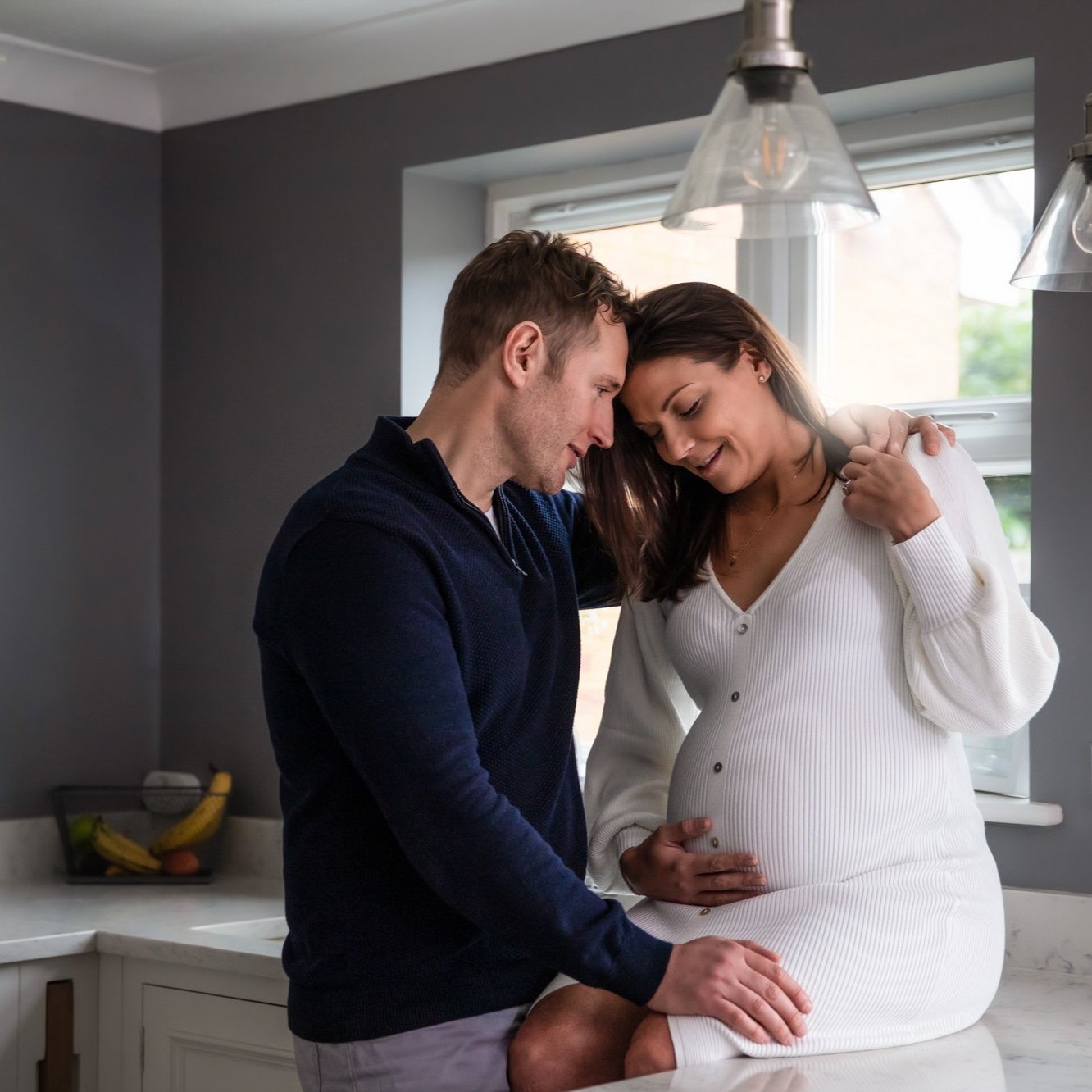 A man and woman share a tender moment in a kitchen, with heads touching and eyes closed, the woman is visibly pregnant, and they are holding her belly gently.