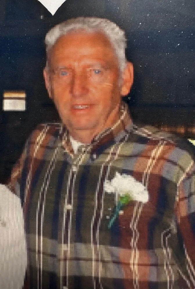Older man with gray hair and blue eyes wearing a plaid shirt and a white boutonniere, indoors with a dark background.