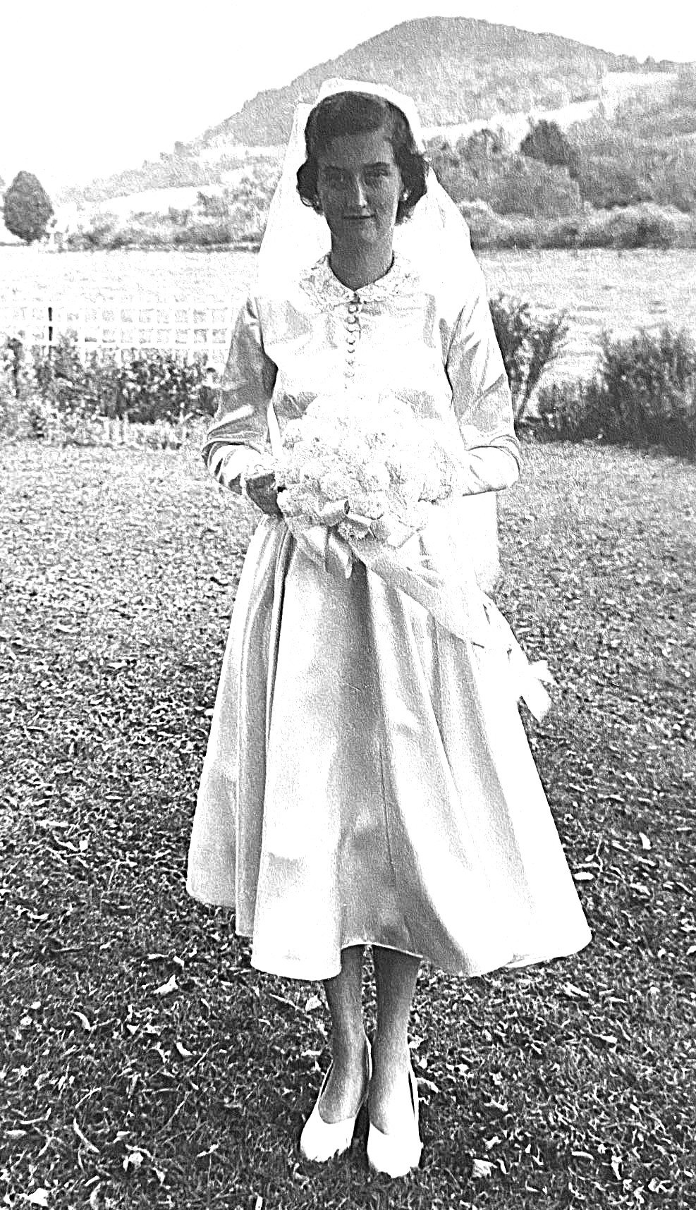 Black and white photo of a woman in a vintage dress holding a bouquet of flowers, standing outdoors with mountains and a fence in the background.