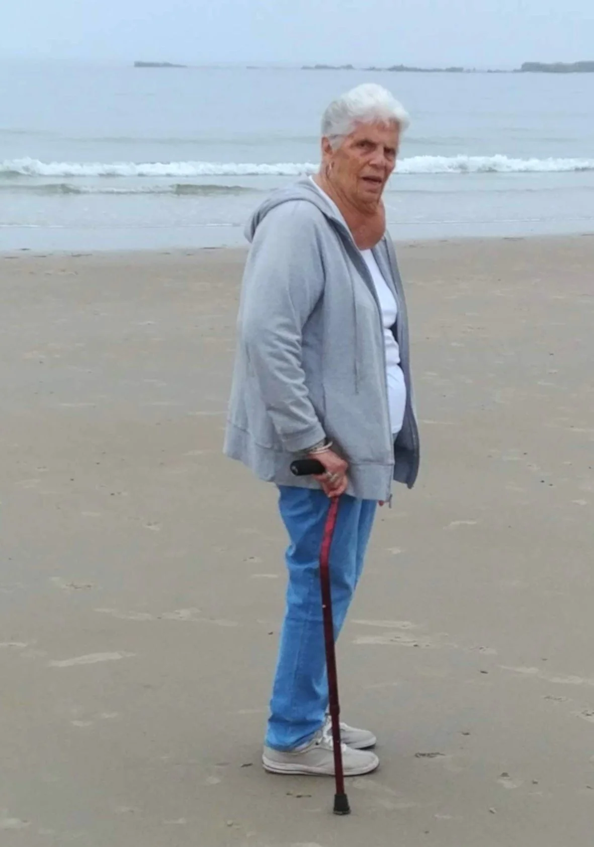 An elderly woman with gray hair, wearing a gray hoodie, white shirt, and blue jeans, stands on a sandy beach near the water, holding a walking cane and a small object in her right hand, with an overcast sky in the background.
