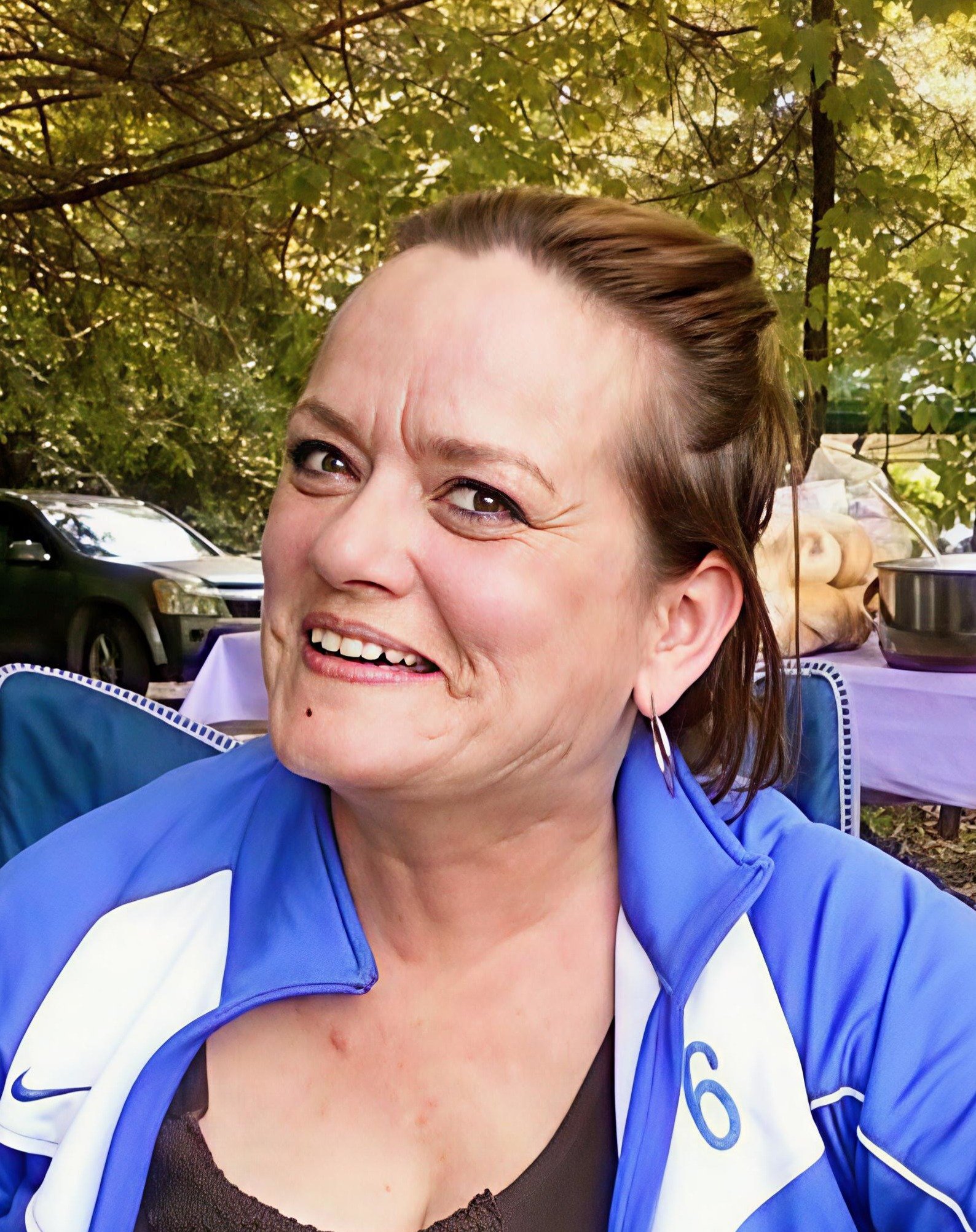 A woman with short brown hair smiling outdoors at a gathering, wearing a blue and white sports jacket, with trees and a table with food and cakes in the background.