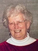 An elderly woman with curly gray hair, wearing glasses, a white turtleneck, and a burgundy sweater, smiling.