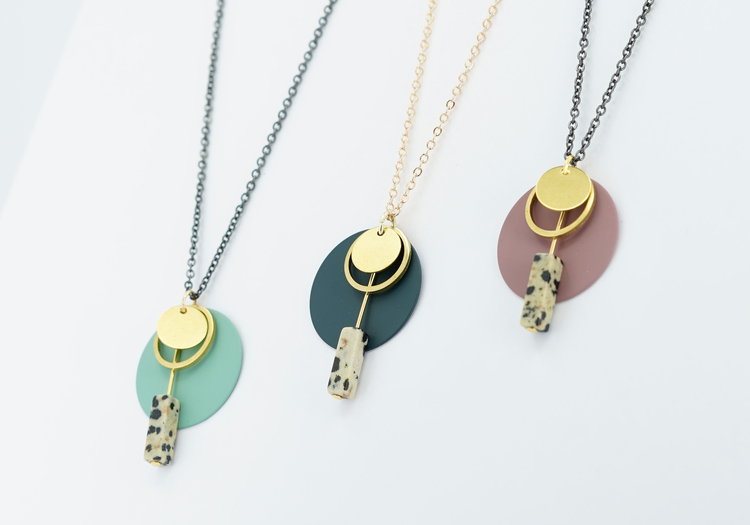 Three modern necklaces with geometric pendants in gold, teal, black, and pink colors.