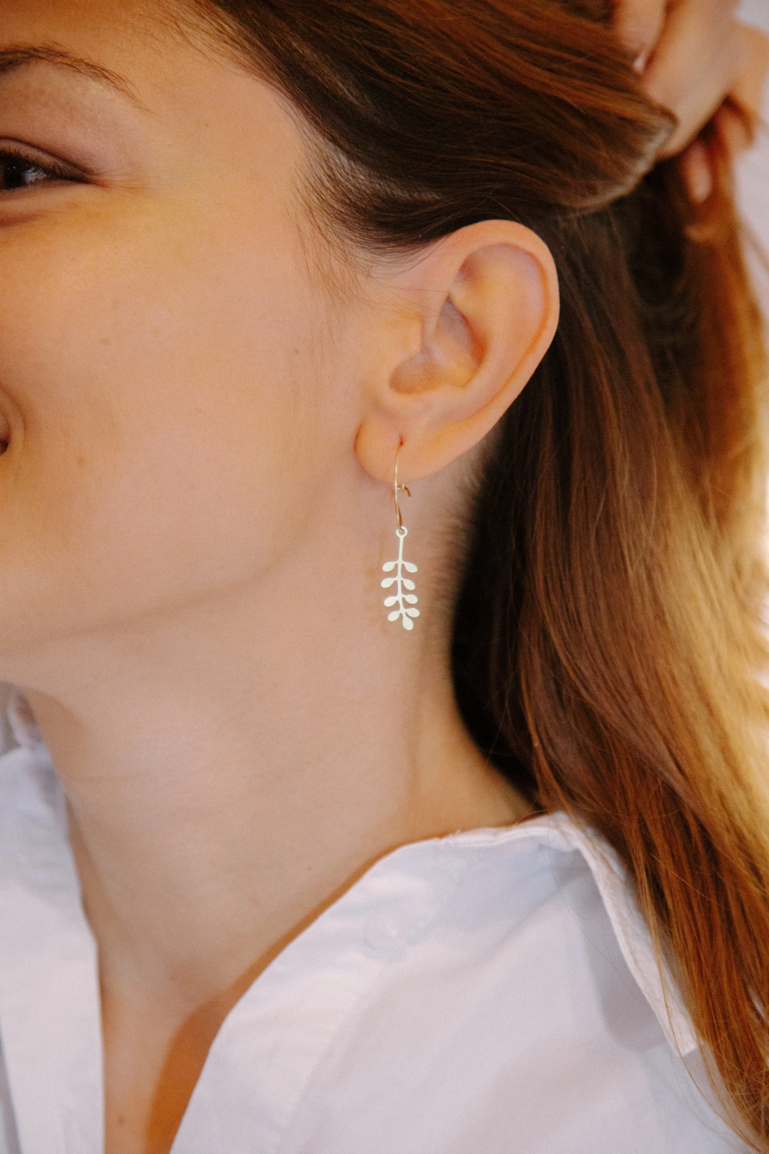 Close-up of a person's ear wearing a dangling leaf-shaped earring, with long brown hair and a white shirt.