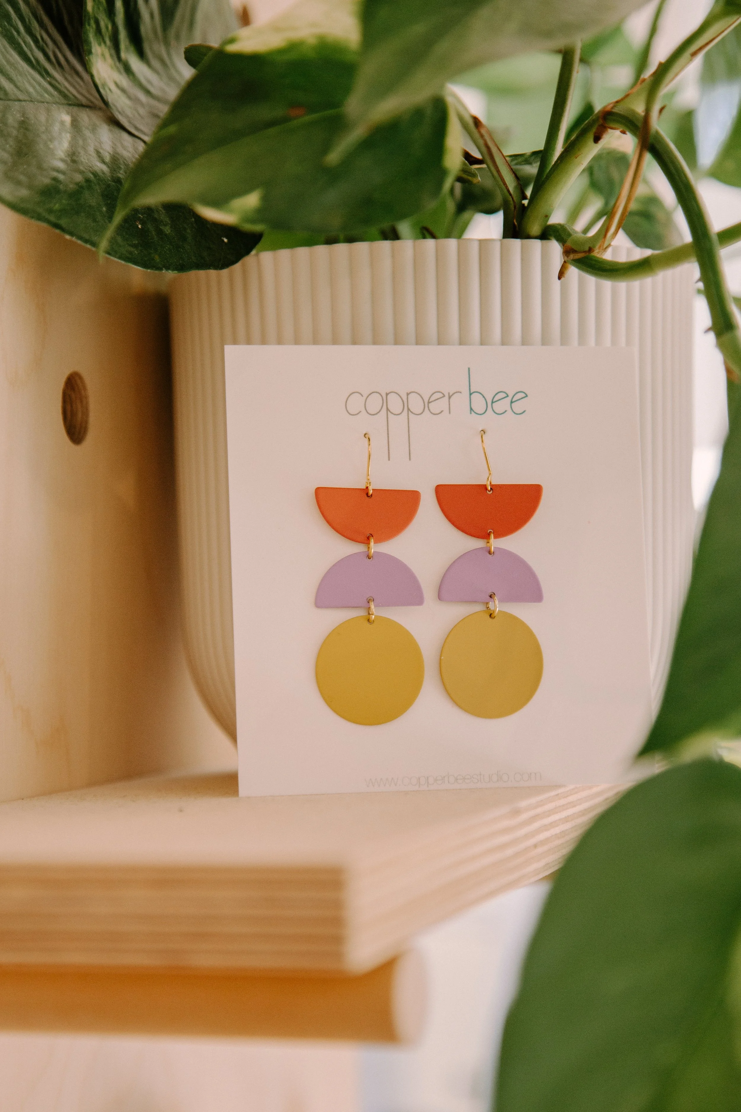 Colorful handmade geometric earrings displayed on a white card with "copper bee" logo, set against a leafy plant in a white pot.