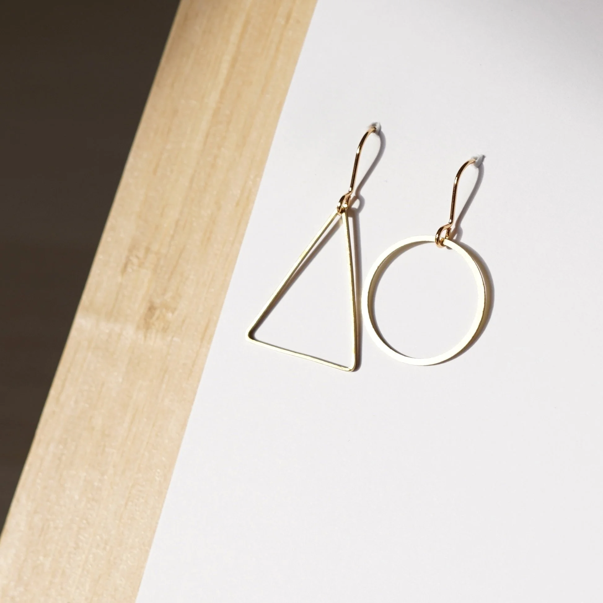 Two geometric-shaped gold earrings, one triangular and one circular, on a white and wooden background.