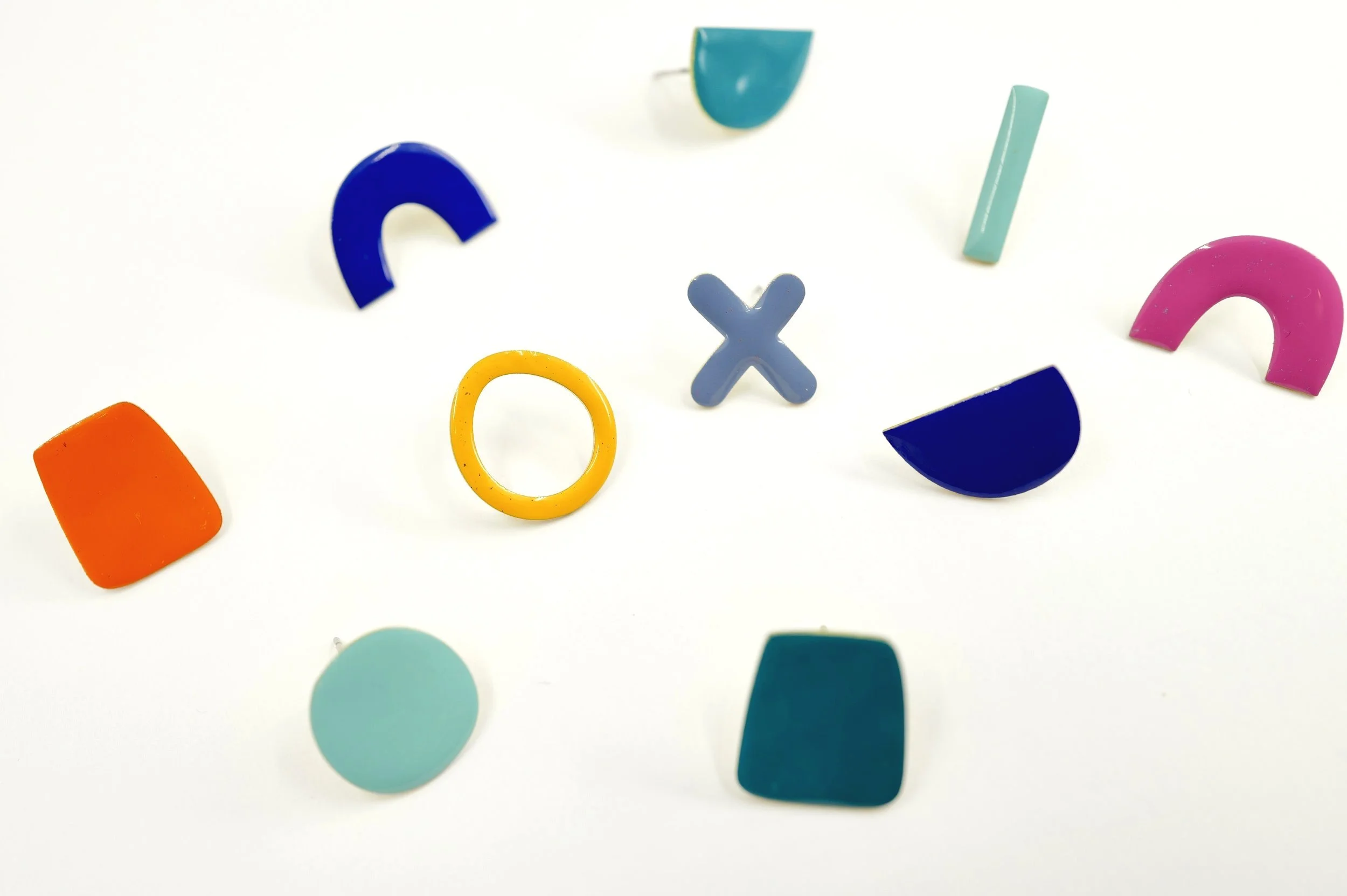 Colorful geometric earrings scattered on a white surface, including shapes like arches, semicircles, and circles in blue, pink, orange, yellow, and teal.