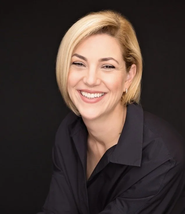 A woman with short blonde hair smiling, wearing a black shirt against a dark background.