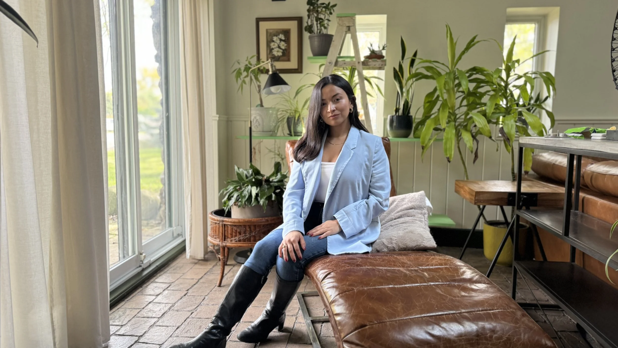 A woman in a light blue blazer and jeans sitting on a brown leather bench in a bright room with large windows, green plants, and wooden furniture.