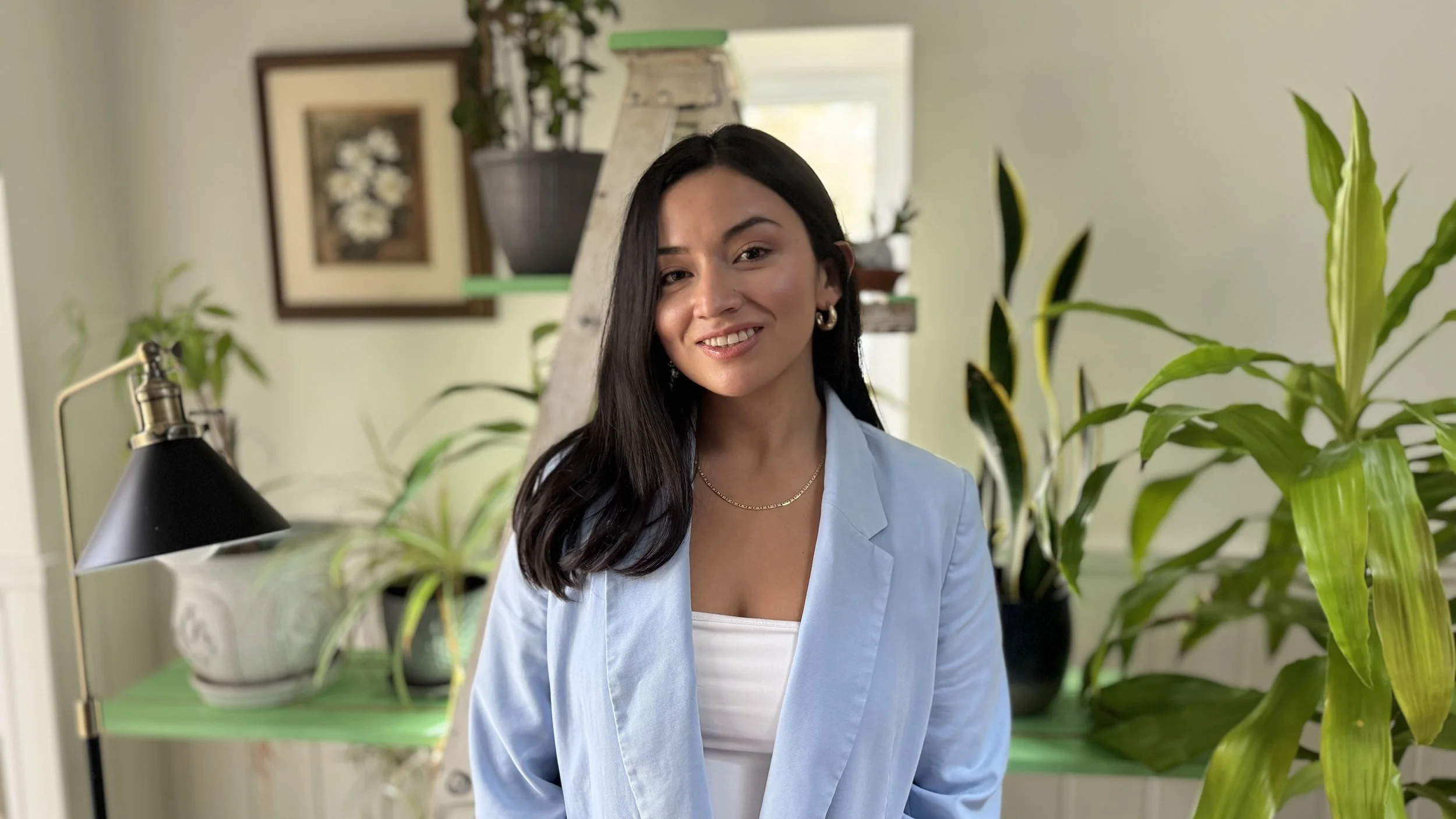 green background, right side snake plants, left side black lamp, middle a portrait photo of a woman with black hair, smiling in a blue blazer with a white shirt
