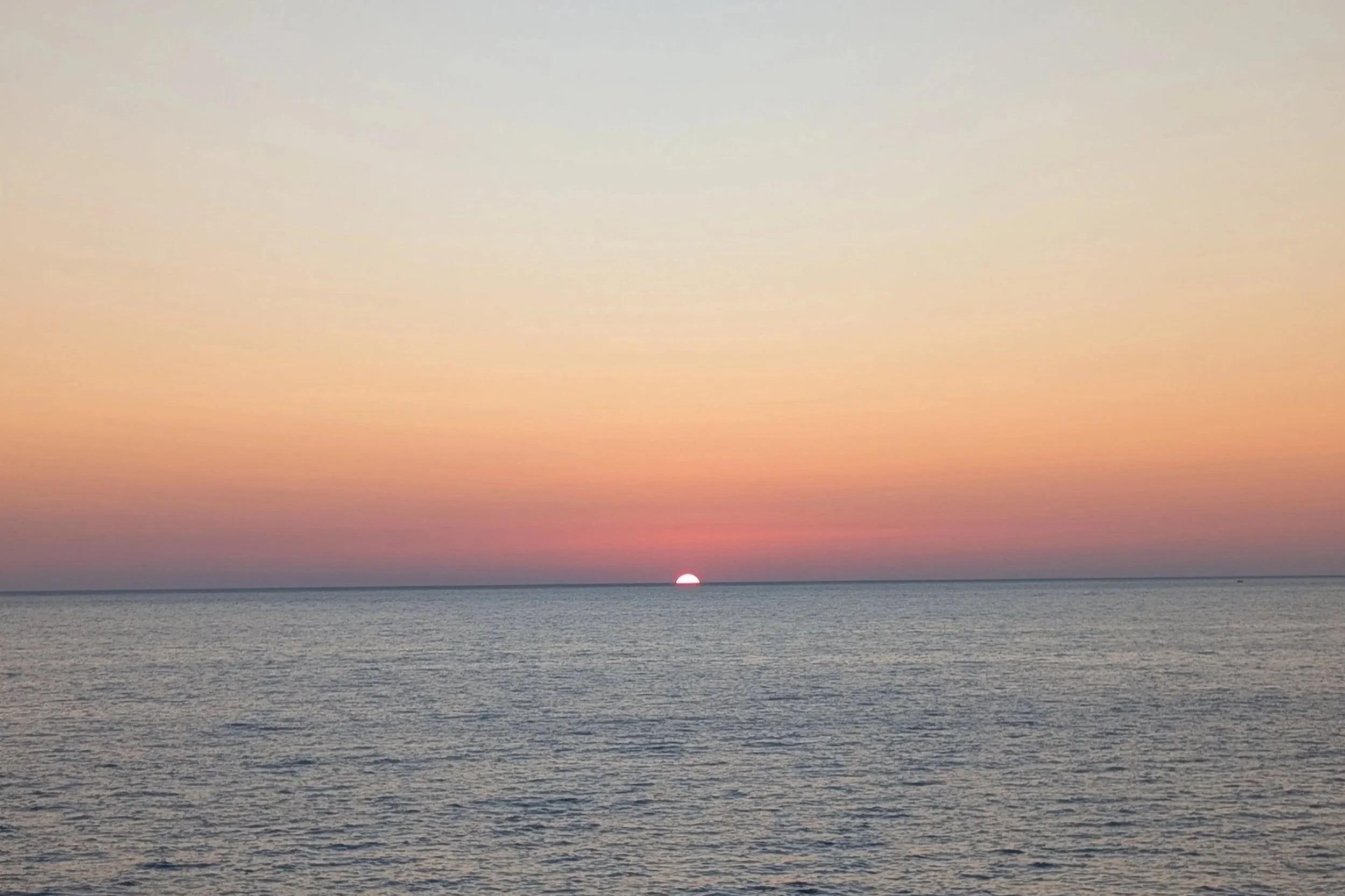 This is an ocean sunset with the sun partially below the horizon, the sky blending pastel pink, orange, and purple hues, and calm water with gentle waves.