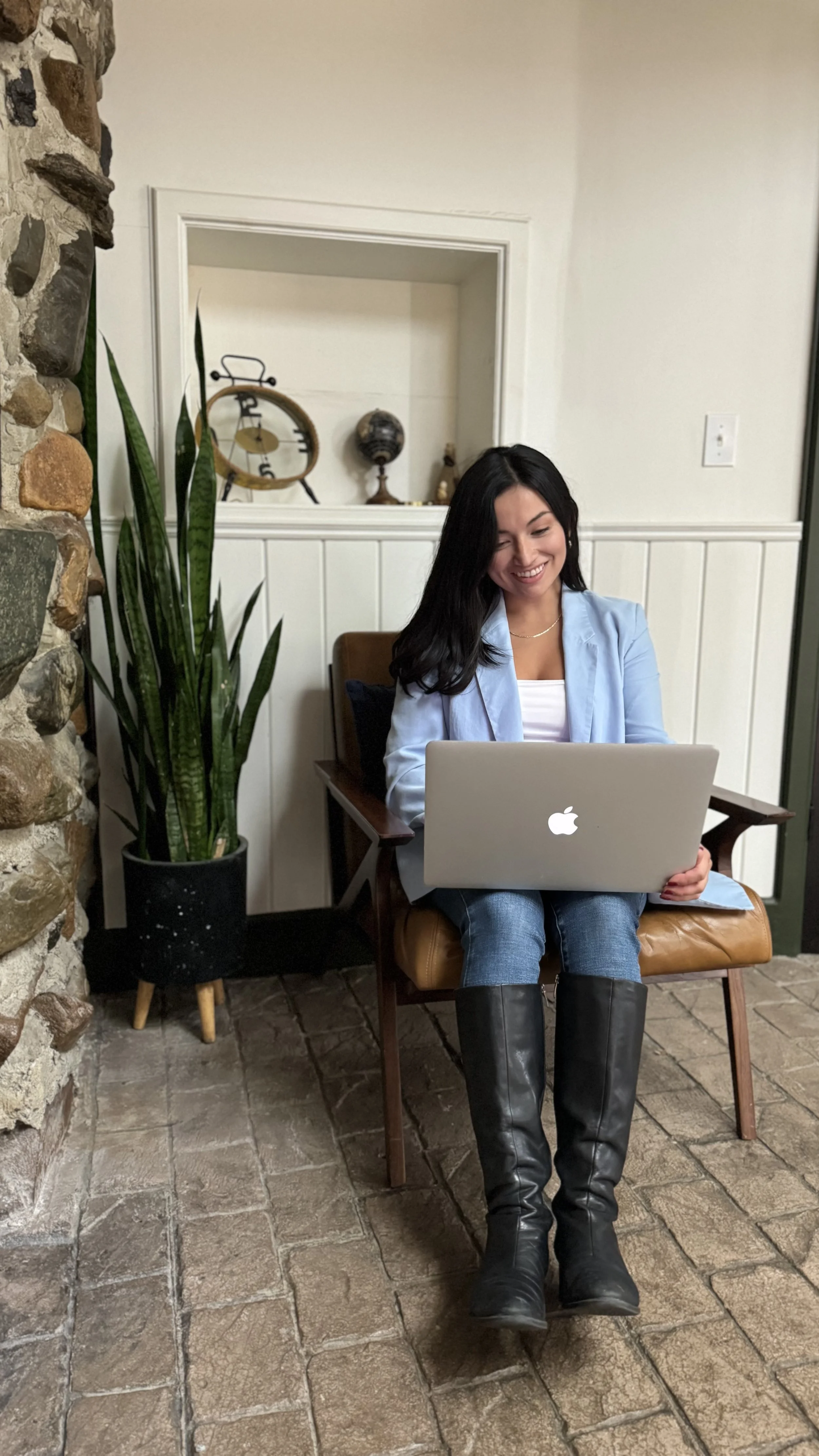 A woman sitting on a brown leather chair with her legs crossed, using a silver MacBook laptop. She has long dark hair and is wearing a light blue blazer, a white top, blue jeans, and black knee-high boots. There is a tall potted plant with long green leaves next to her, and a stone wall partially visible on the left. In the background, there is a decorative wall opening with a vintage clock, a globe, and other decorative items.
