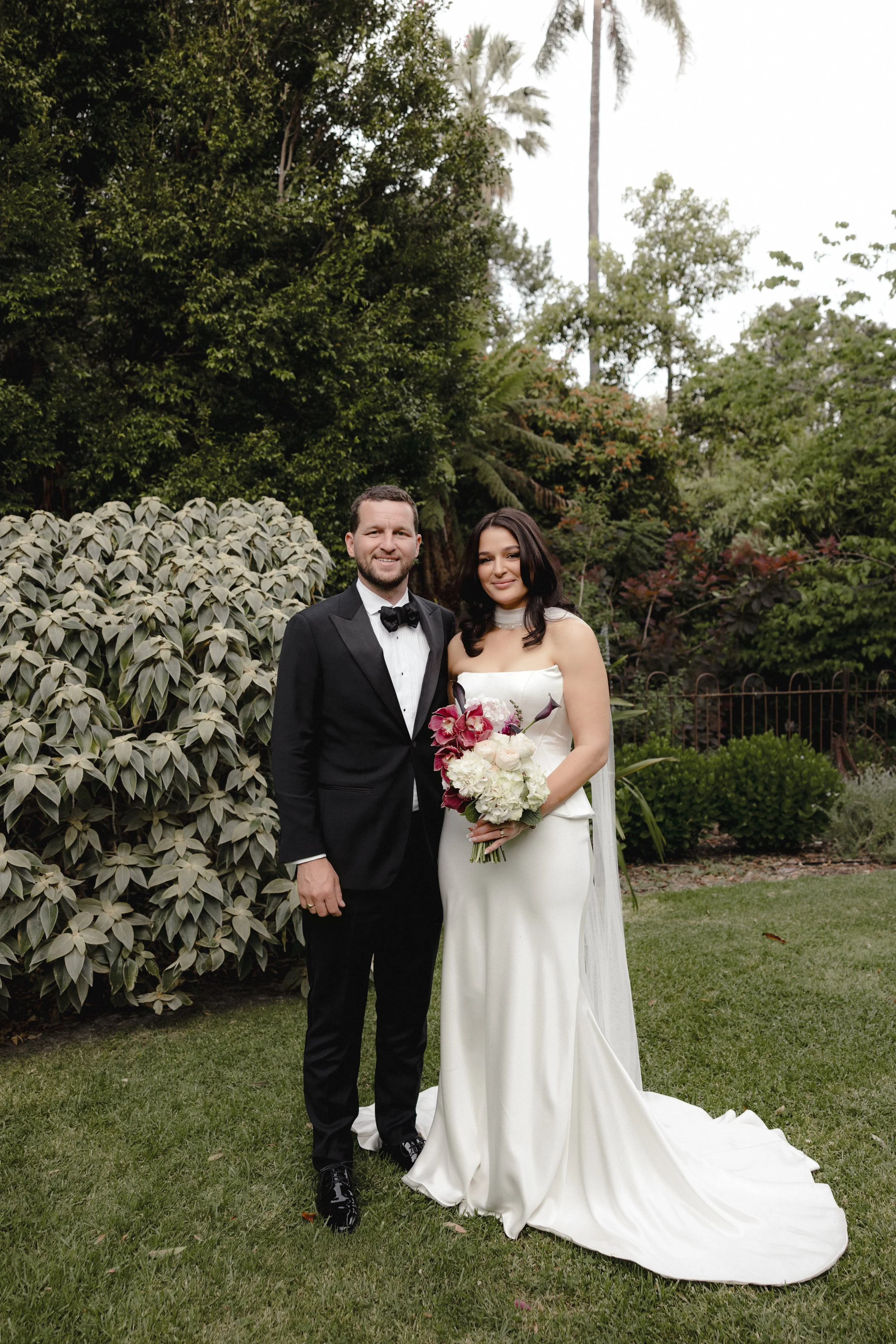 Newly wed husband and wife stand in the Botanical Gardens at Gardens House with flower bouquet created by Petal and Sage