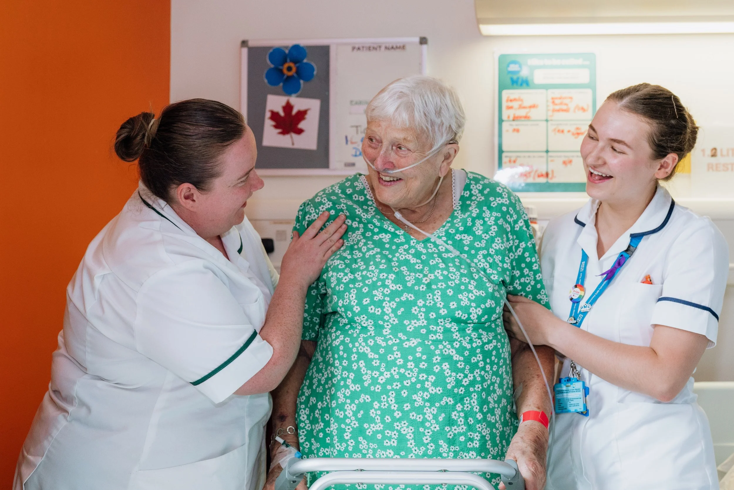A lady in hospital being helped out of her chair by 2 healthcare workers. They are all smiling at her achievement