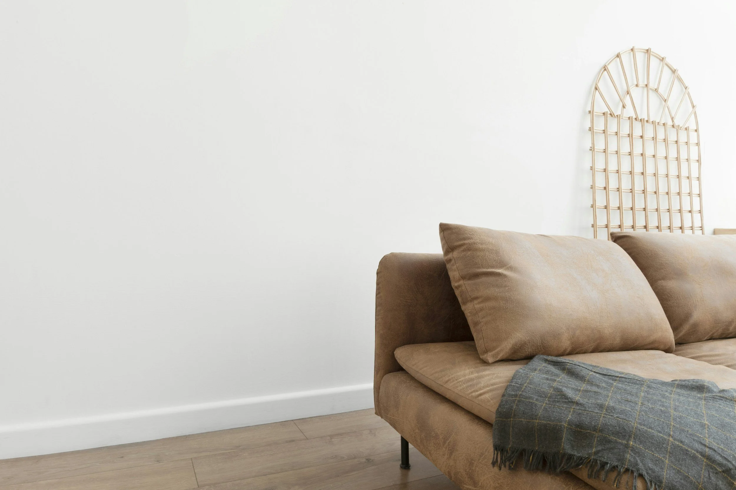 Living room corner with beige sofa, throw blanket, wooden floor, white wall, and decorative headboard.