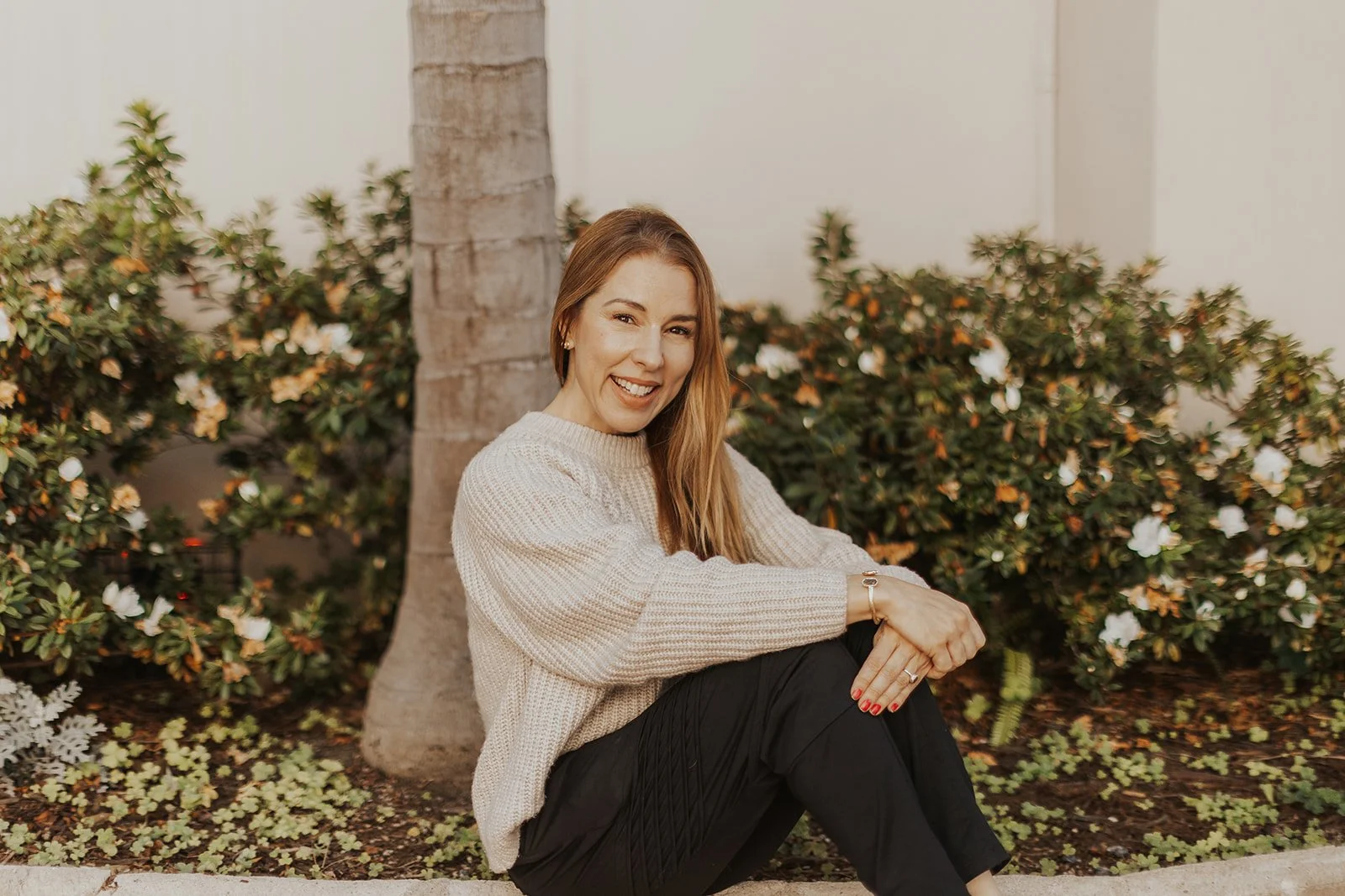 A woman sitting outdoors next to a palm tree, smiling at the camera, with bushes in the background.