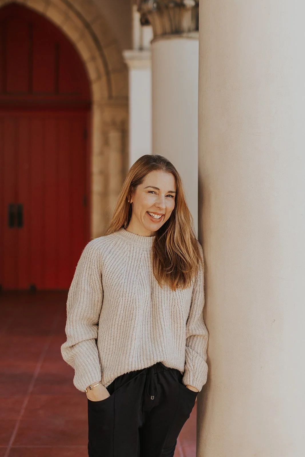 A smiling woman in a beige sweater and black pants standing next to a large column outside a building with red doors.
