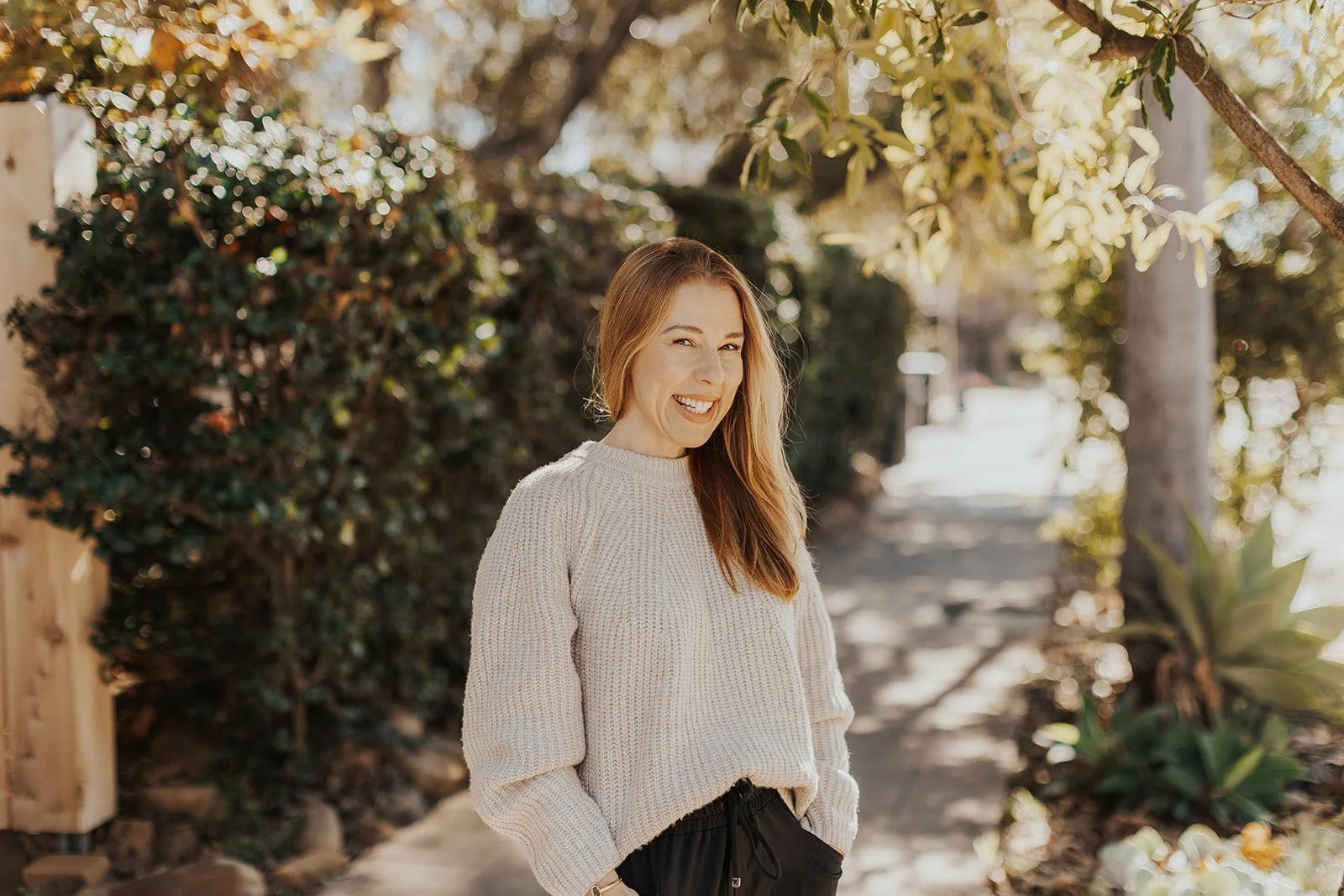 A woman with long red hair smiling outdoors on a sunny day, wearing a cream knit sweater and black pants, standing on a wooded path with trees and plants around.