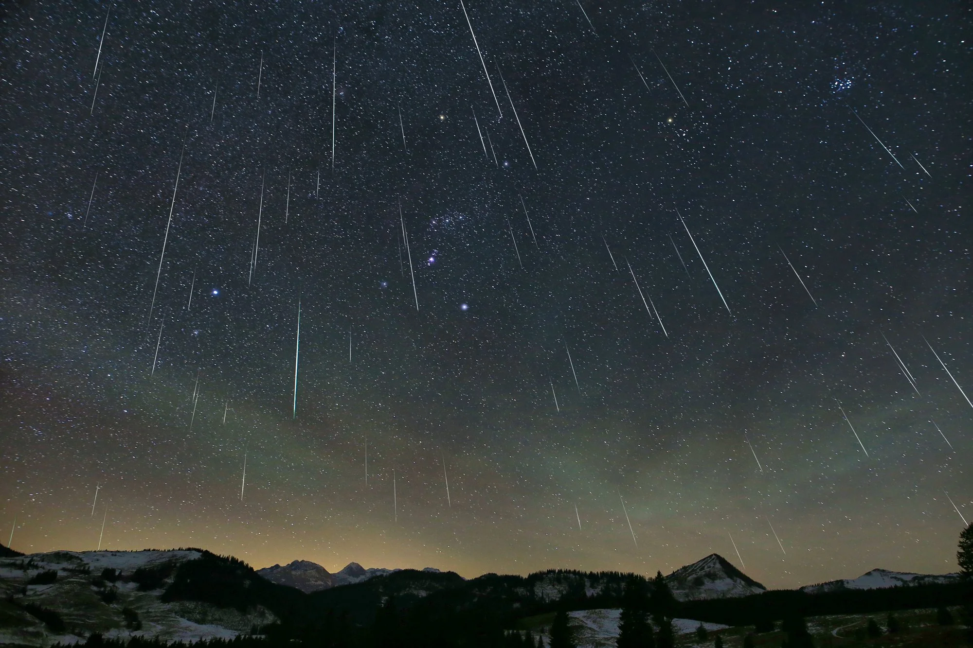 Composite image of the Geminid meteor shower above snow covered mountains at Postalm, Austria, with dozens of bright meteor streaks crossing a star filled sky that includes Orion and the Pleiades.