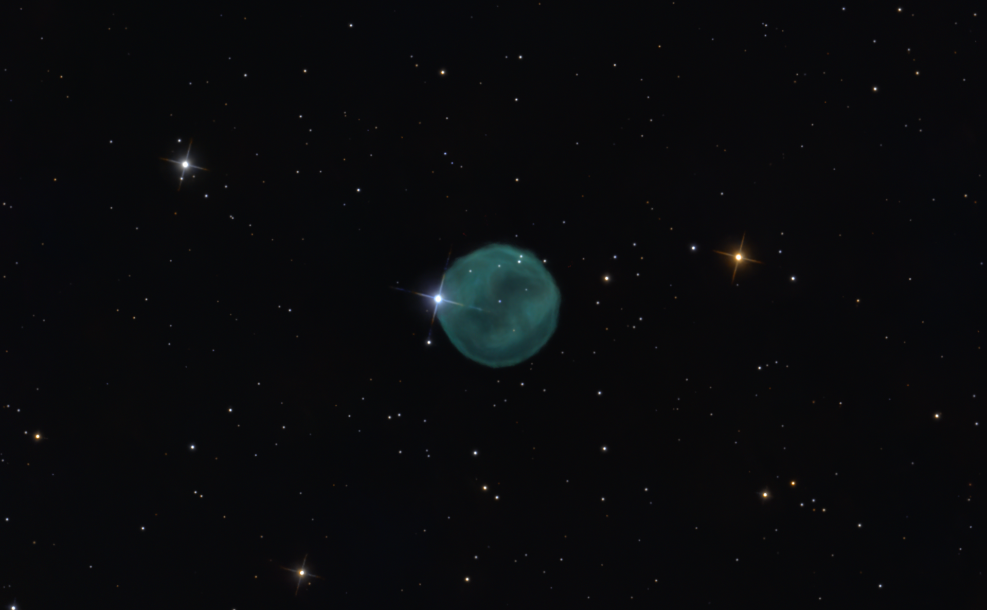 A faint turquoise spherical planetary nebula with a bright star overlapping its edge, creating a diamond ring appearance against a dense star field.
