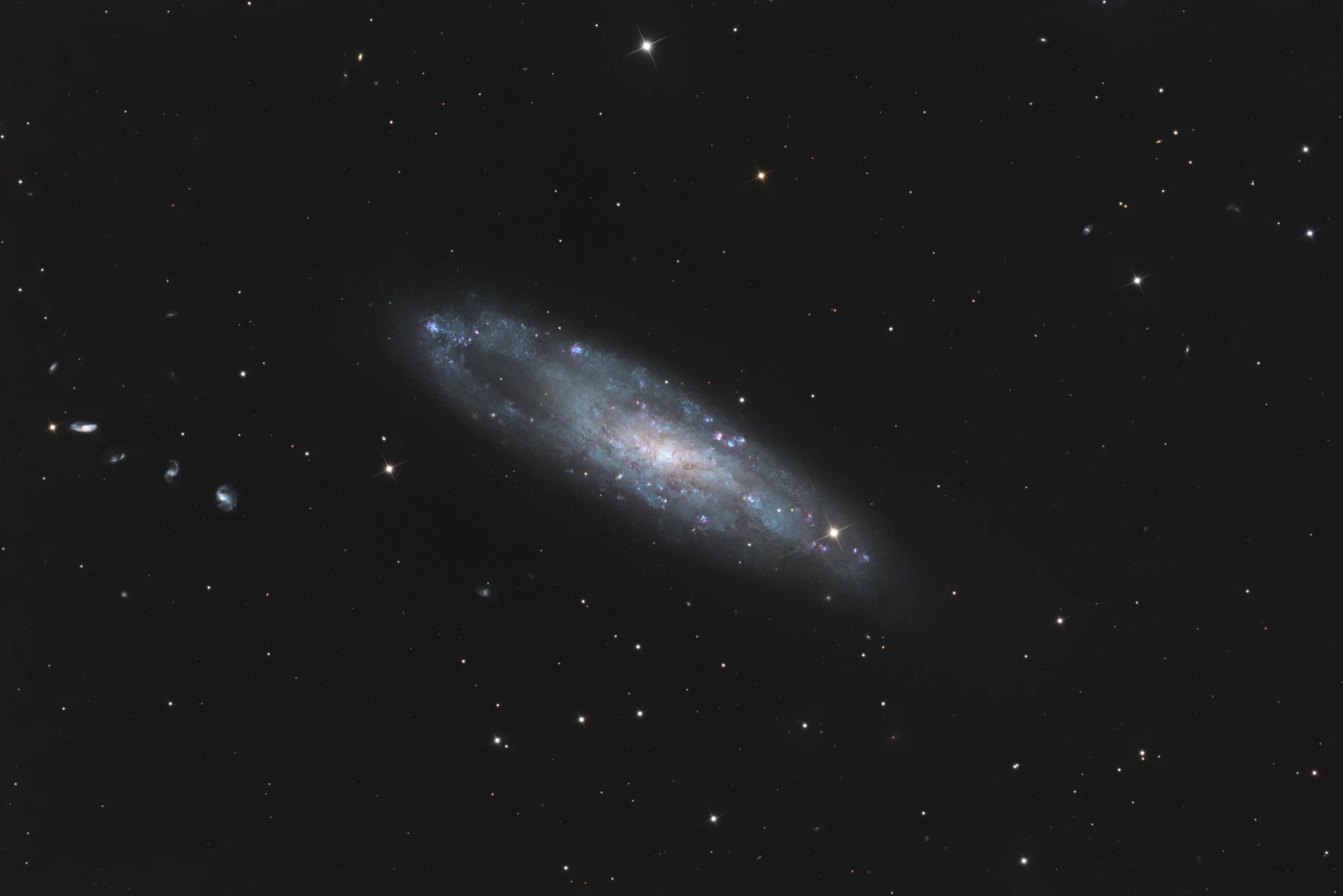 Caldwell 62, NGC 247, an elongated spiral galaxy with faint arms and blue star-forming regions, set against a dark sky scattered with stars and distant background galaxies.